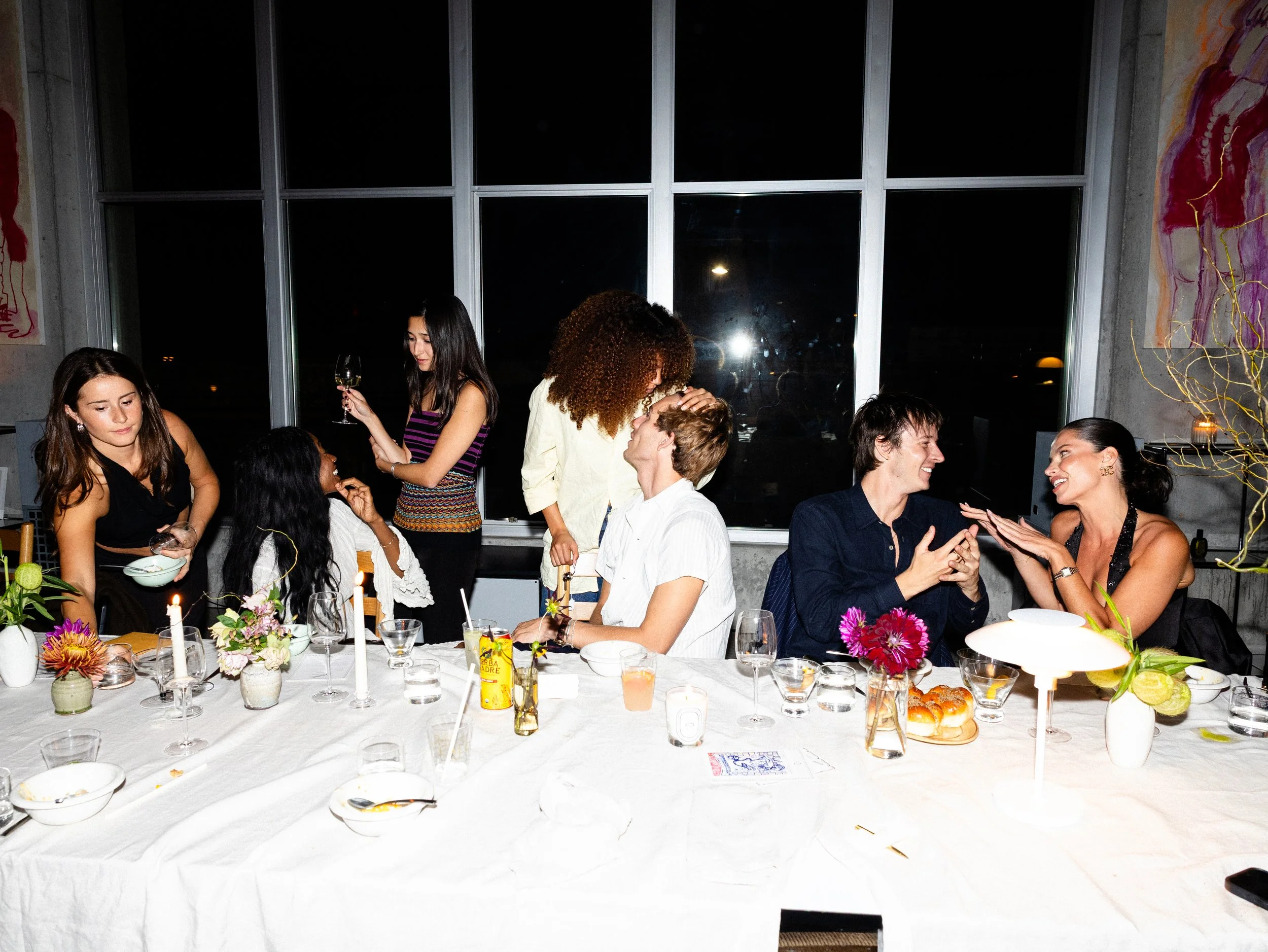 A group of people celebrating at a dinner party around a long table with flowers, candles, and food. Some are sitting and talking while others are standing and smiling.