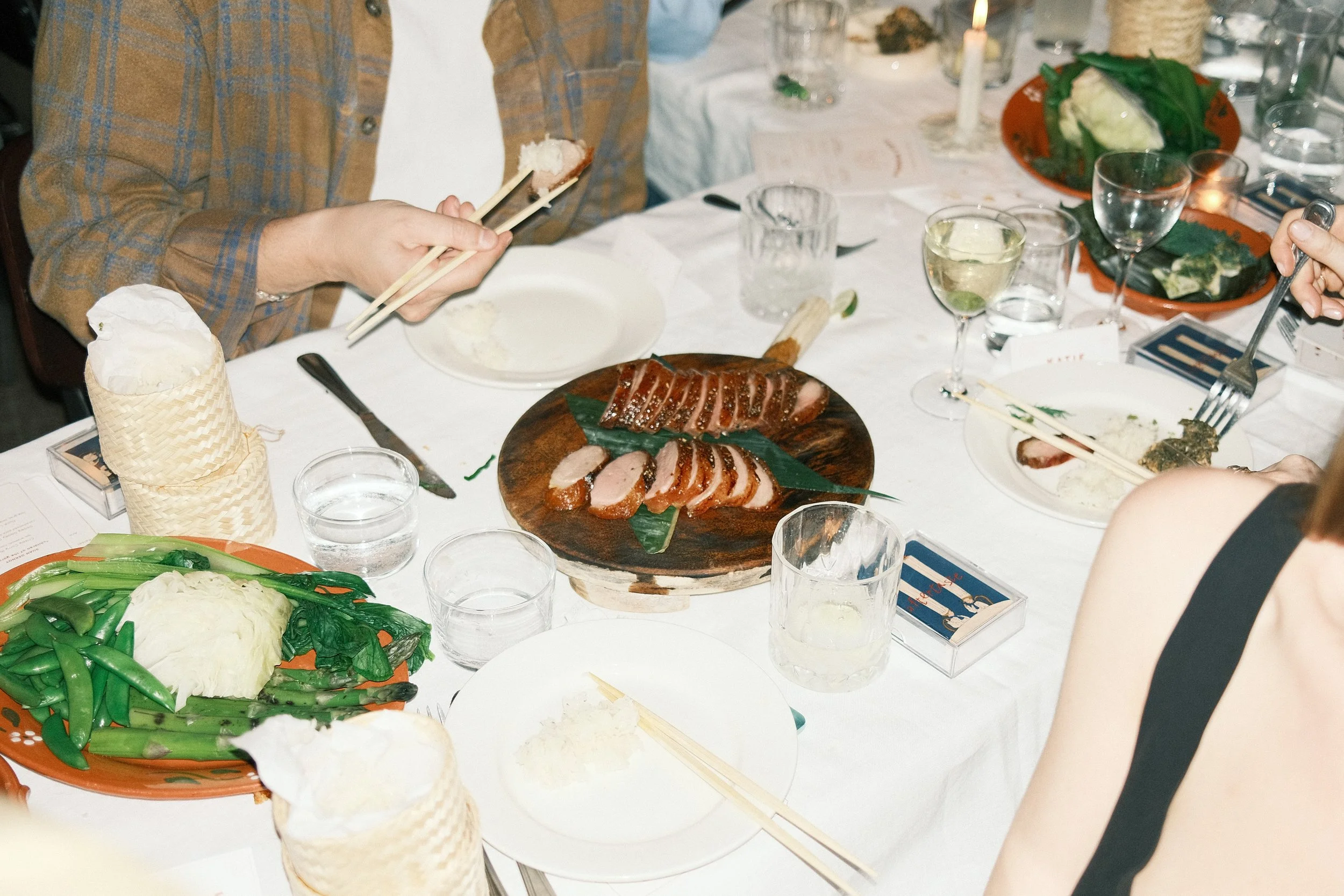 A dinner table with various dishes, including sliced meat, vegetables, and drinks, set for a meal with people serving and eating.