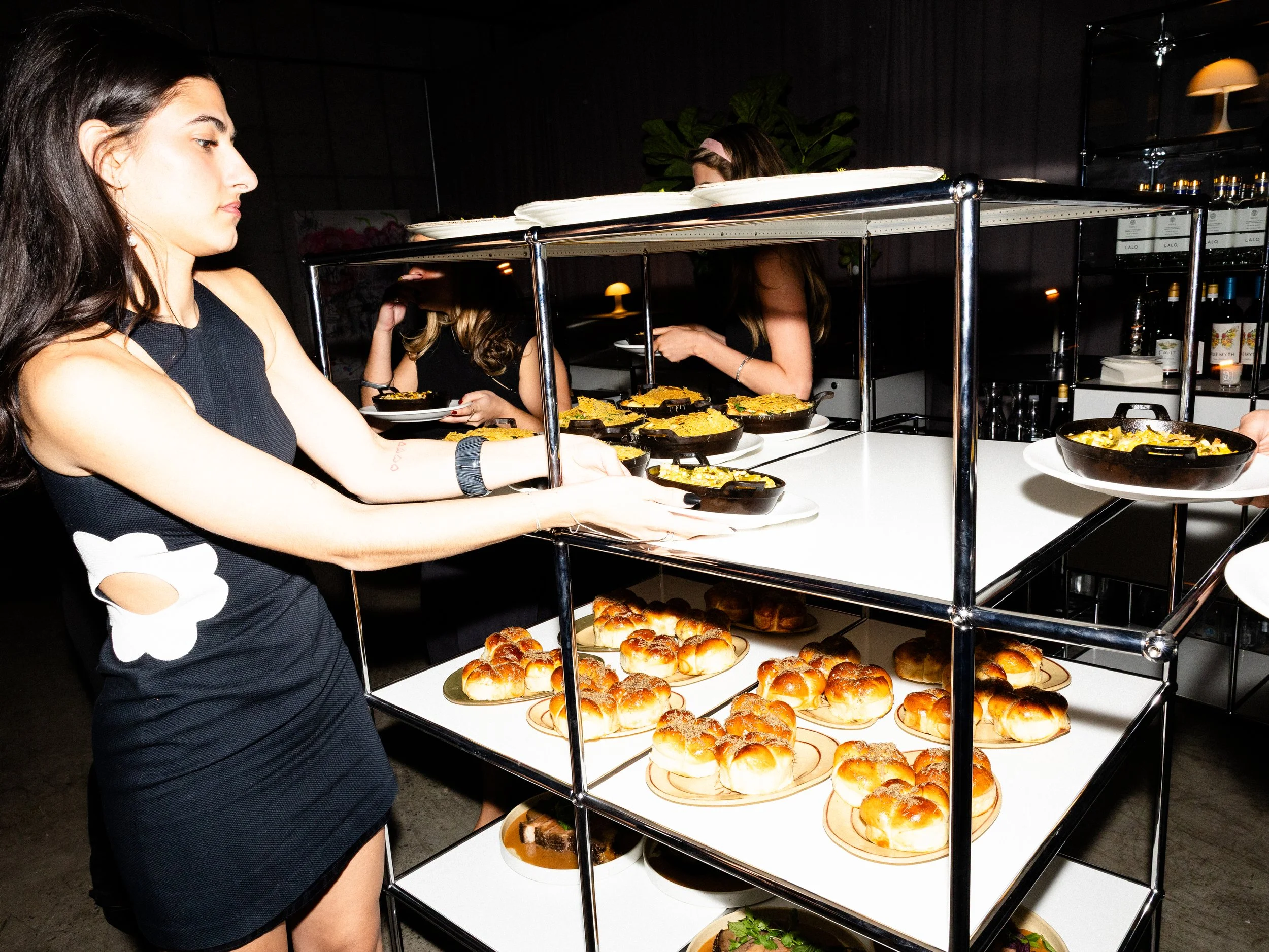 A woman at a buffet table with a glass cover, serving herself food. The table has various dishes including baked goods and pasta under the glass cover and on plates.