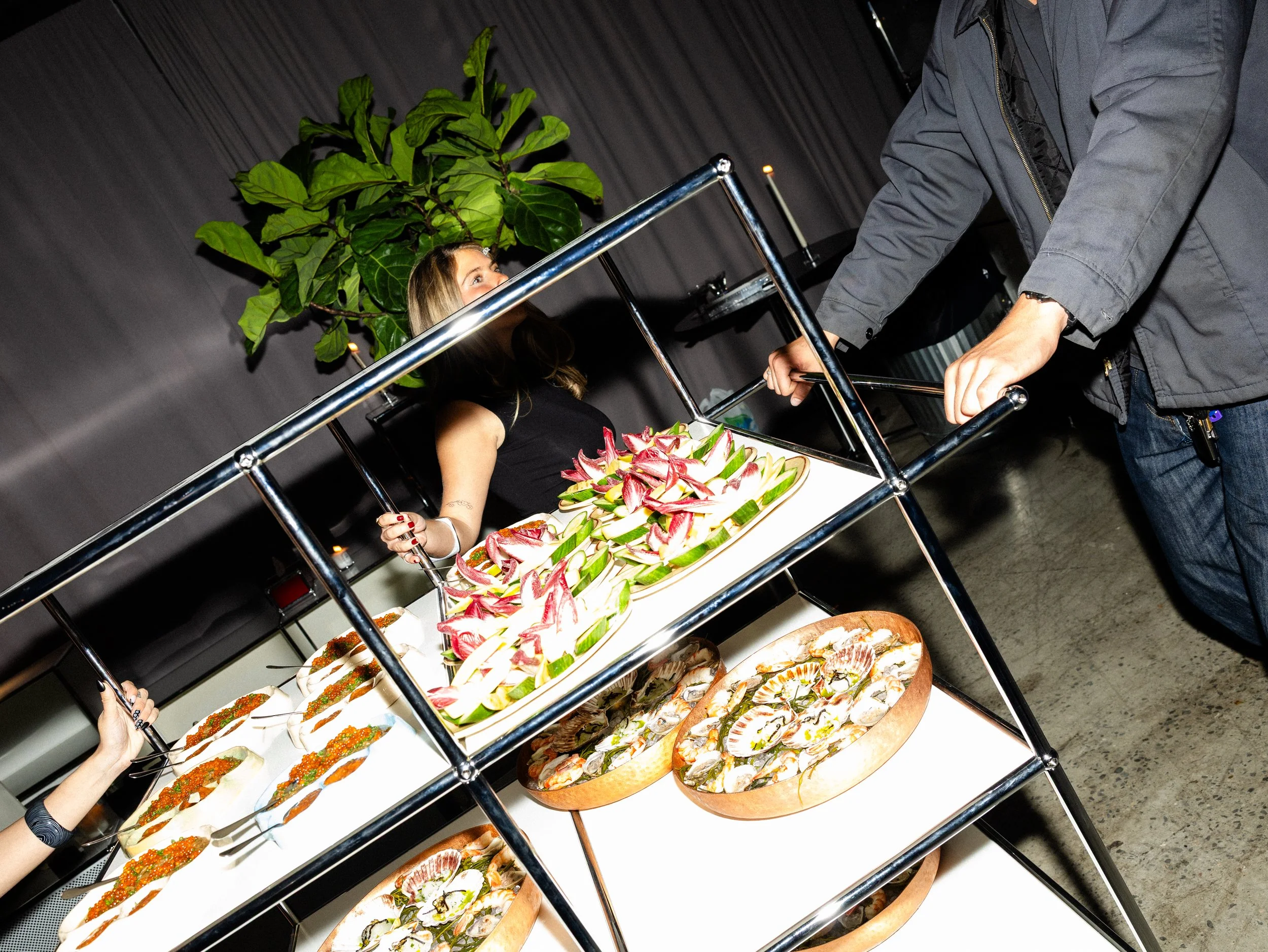 A woman in black holding a serving fork stands behind a food cart with various plates of fish and seafood sashimi, vegetables, and garnishes on display.