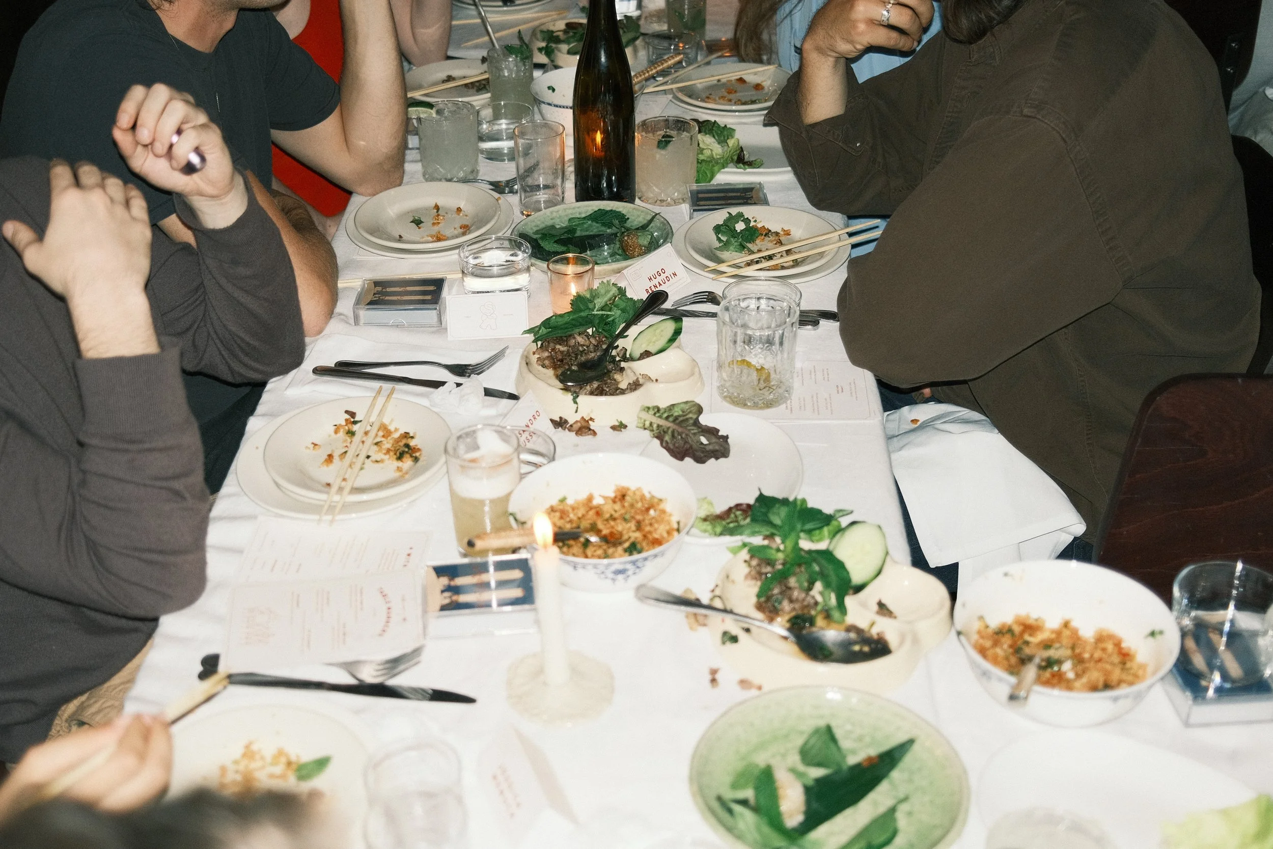 A dinner table with partially eaten plates of food, glasses of water and drinks, chopsticks, and a variety of dishes including salads and rice. Several people are seated around the table, some with their hands resting near their plates.