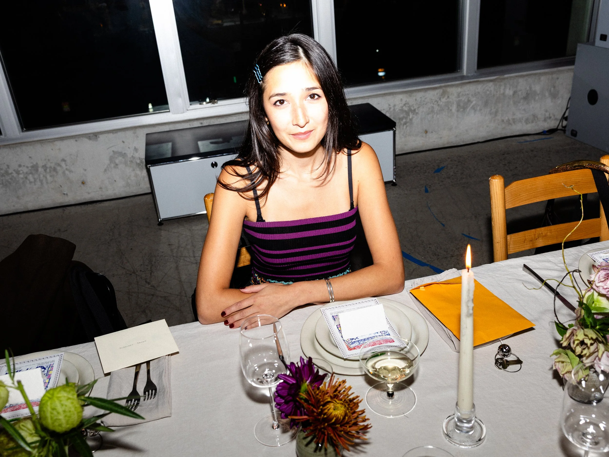 A young woman with dark hair, wearing a purple and black striped sleeveless top, sitting at a dining table with a candle, flowers, and name cards, inside a room with a large window at night.