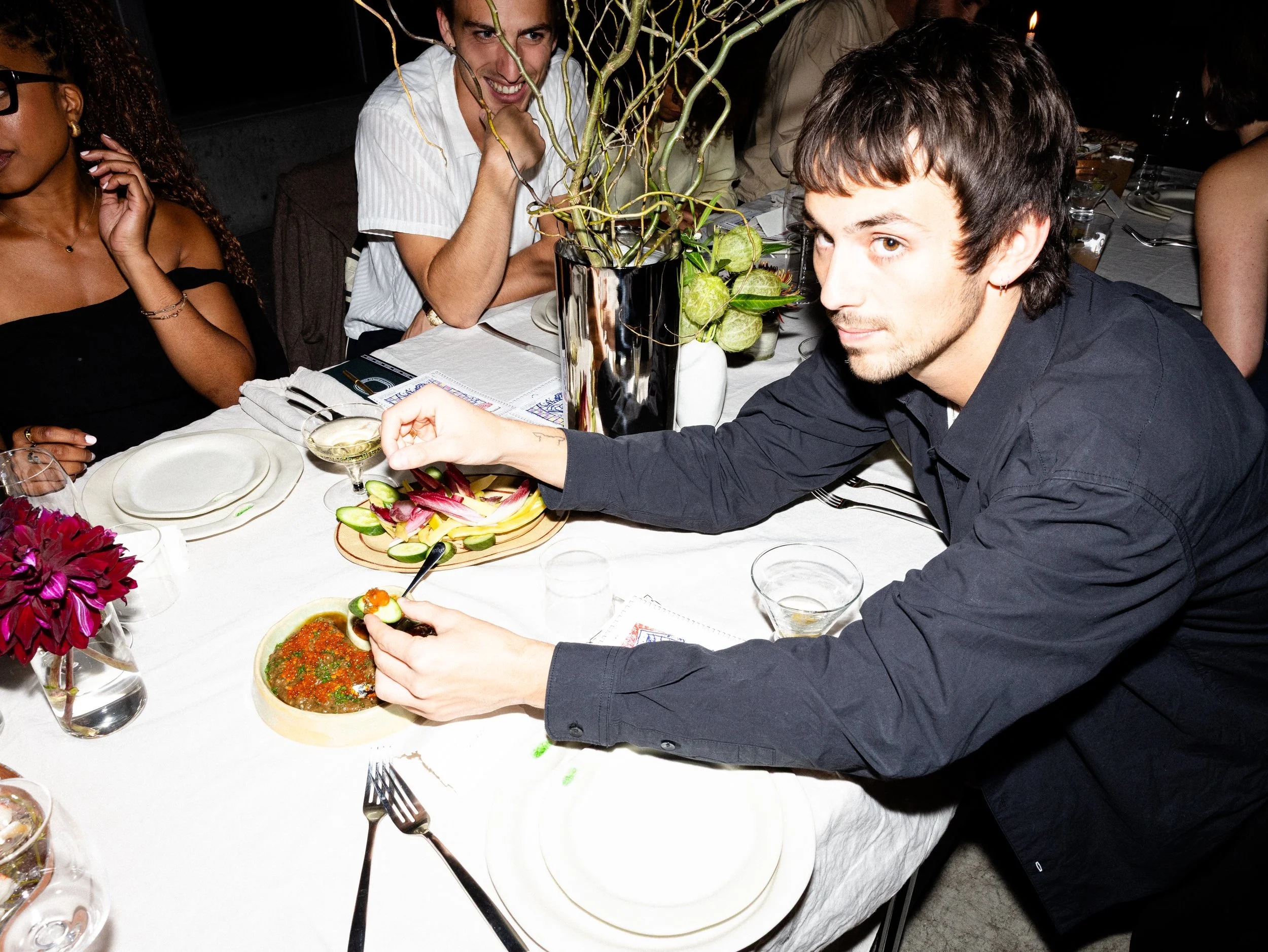 People gathered around a dining table with food and flowers, engaging in conversation and enjoying a meal at a social gathering.