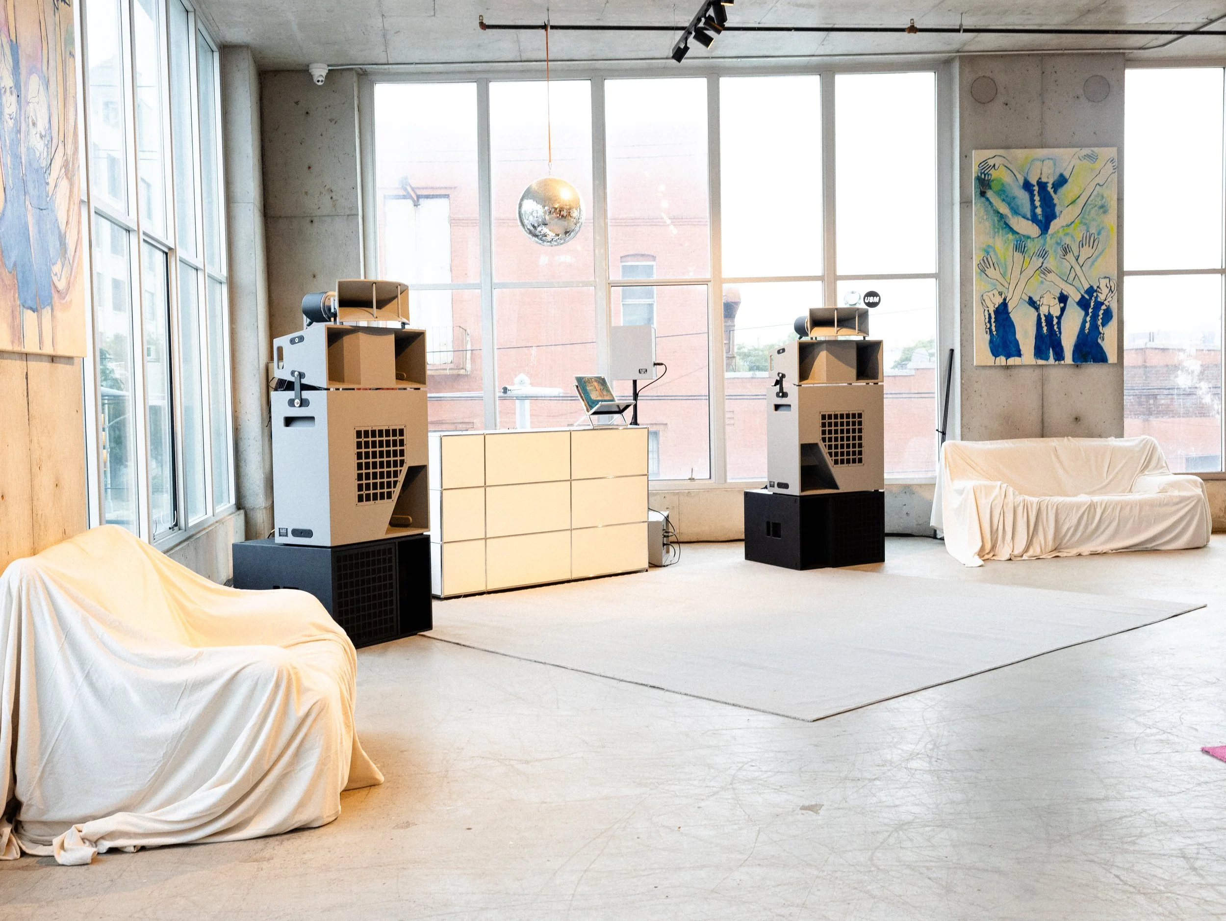 Interior of an art gallery with large windows, modern lighting, abstract artwork on the walls, and furniture covered with white sheets.