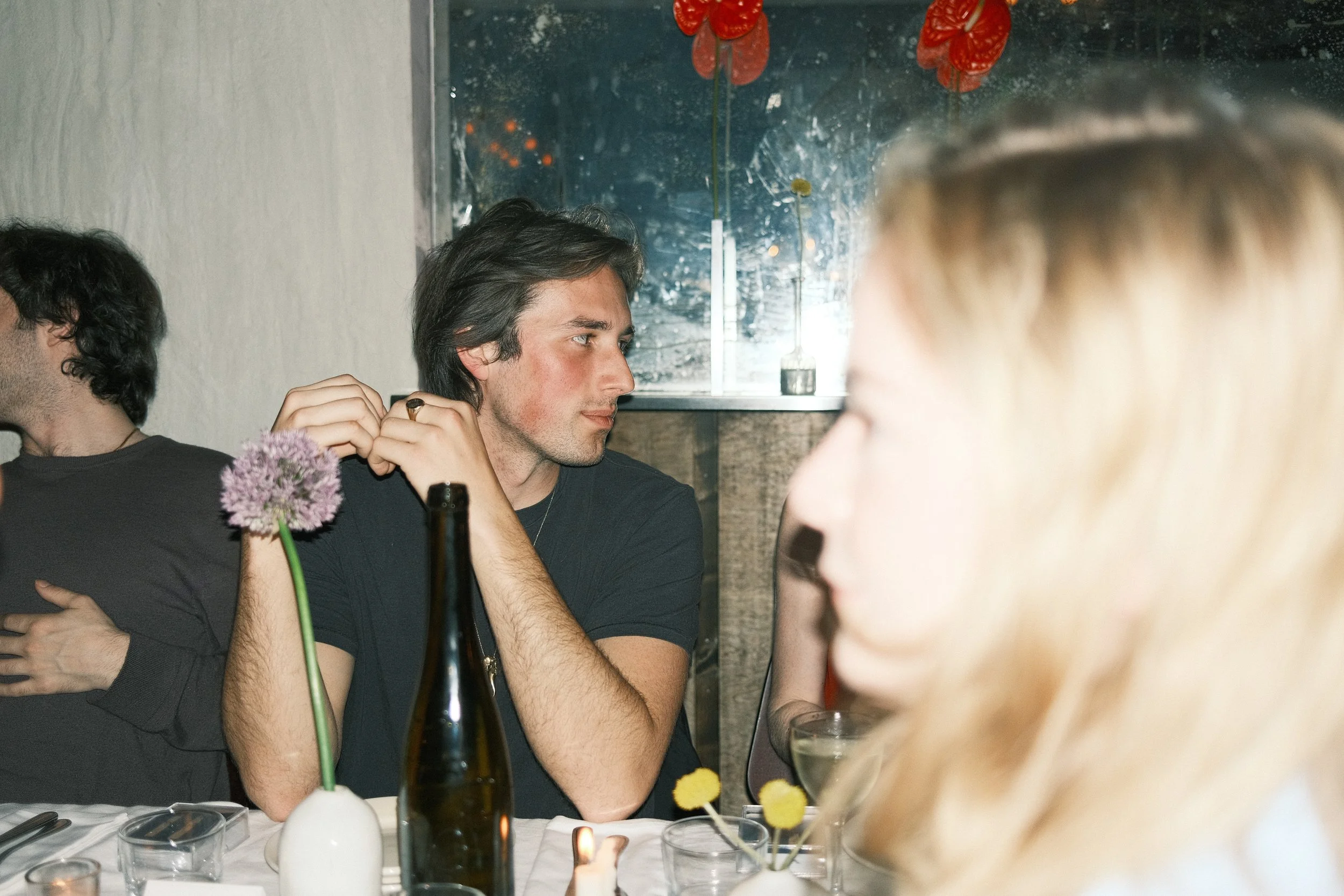 Young man with dark hair and light skin sitting at a dining table, looking to his left. The table has glasses, bottles, and flower arrangements. Other people are partially visible around the table, and a sparkler is burning in front of him.