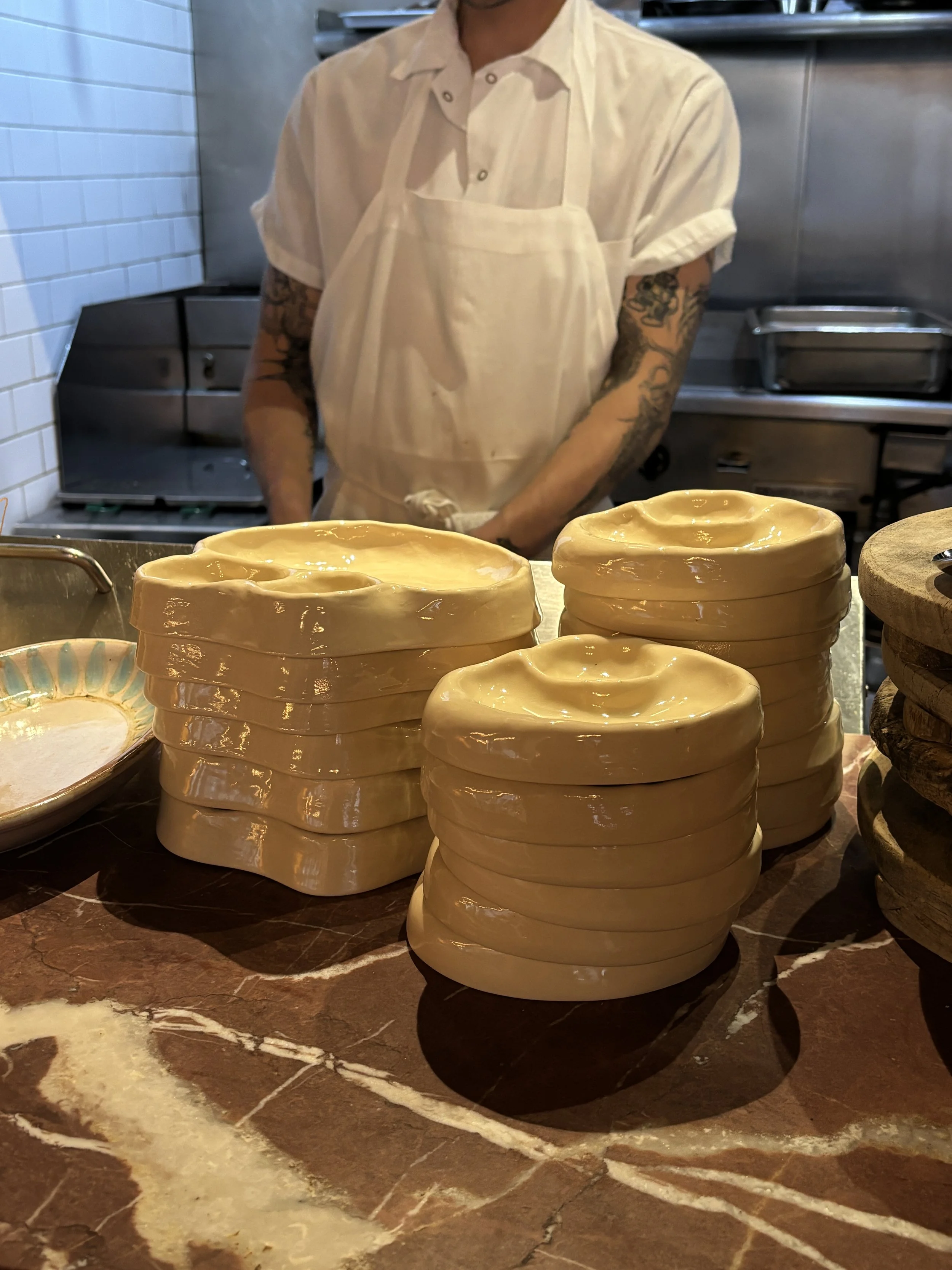 Stacks of yellow cheese rounds on a countertop, with a person wearing a white apron and shirt in the background.