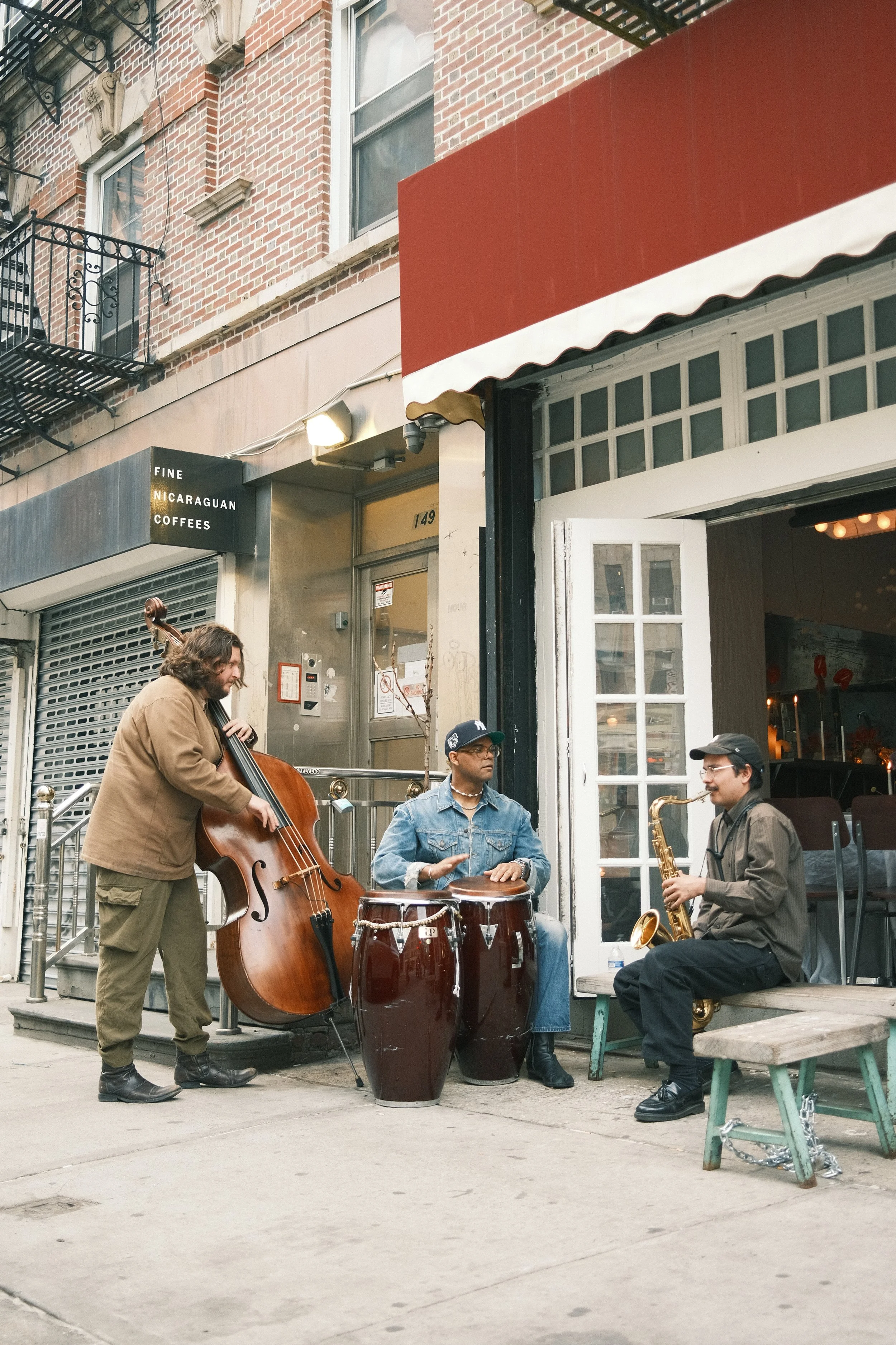 A jazz band performing outside a restaurant on a city sidewalk. The band includes a double bass player, a percussionist, and a saxophonist. The restaurant has a red awning and a partially open white door, with a brick building and window in the background.