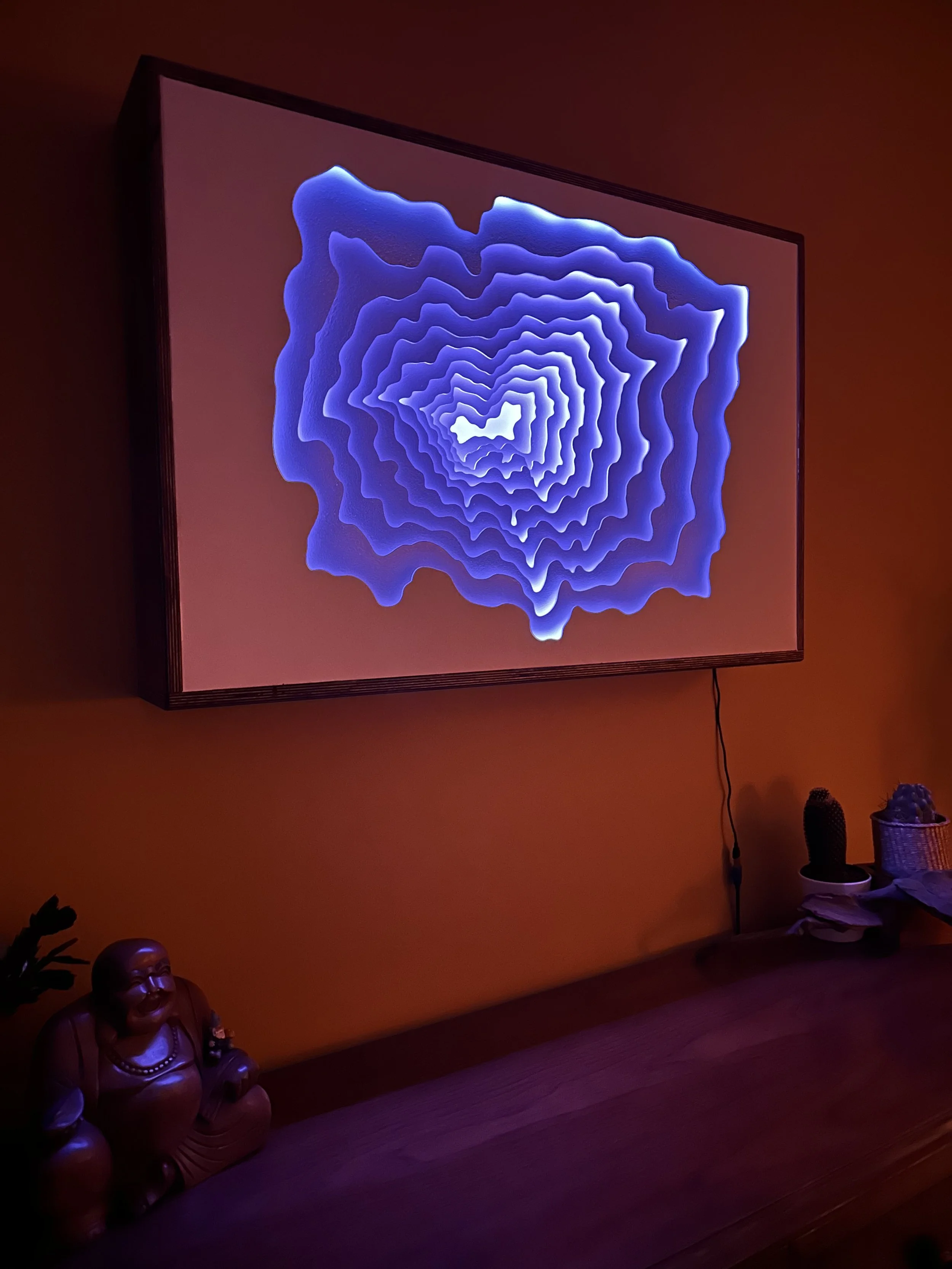 A digital art display on a wall shows a blue, concentric, layered, abstract shape resembling a tunnel or vortex against a reddish background, with a wooden surface below holding a decorative figurine, a potted plant, and some miscellaneous objects.