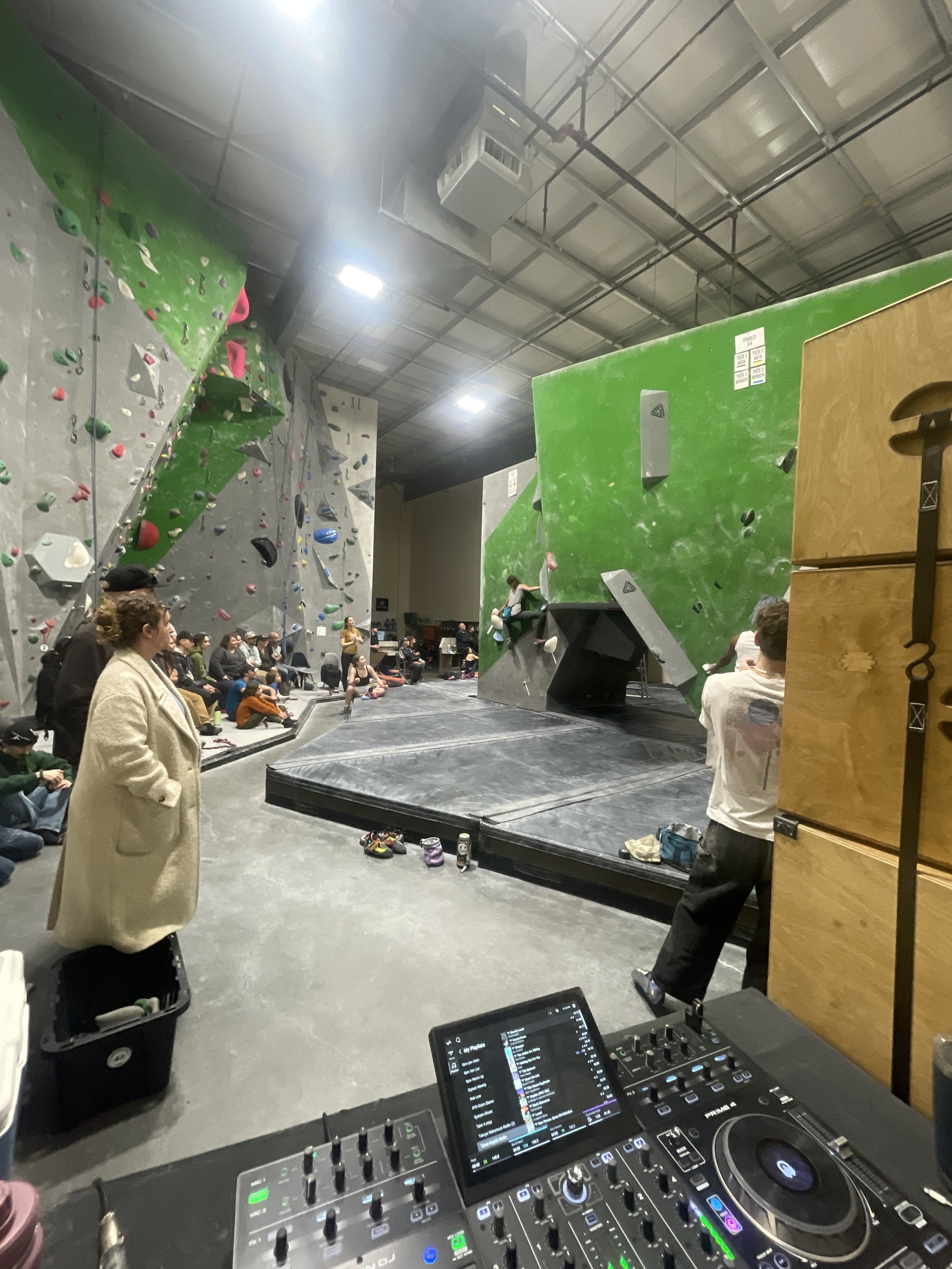 Indoor rock climbing gym with people climbing on various colored walls, spectators seated, and DJ equipment set up in the foreground.
