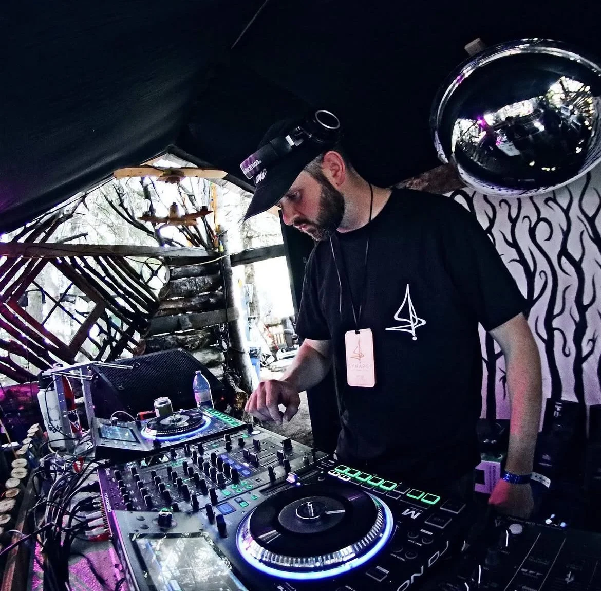 A DJ wearing a black T-shirt and a black cap is playing music with a DJ controller under a black canopy outdoors, with trees visible in the background.