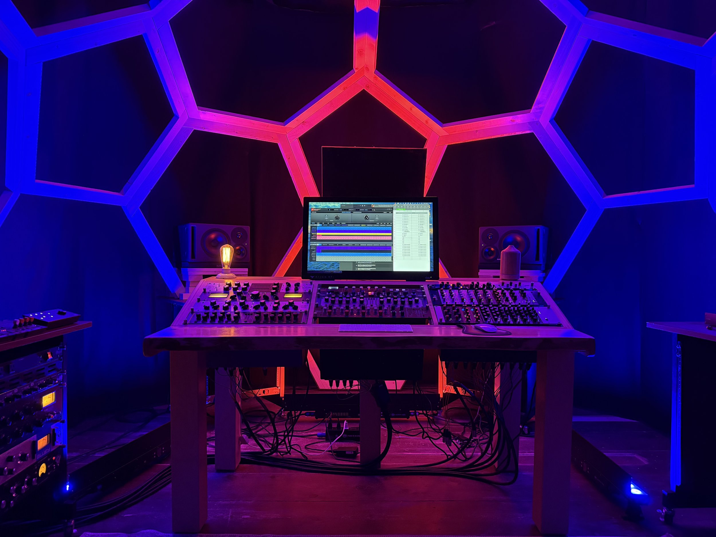 Music production studio with a large mixing console, a computer displaying digital audio workstation software, studio monitor speakers, LED lighting, and musical equipment in a dark room with blue and purple lighting.