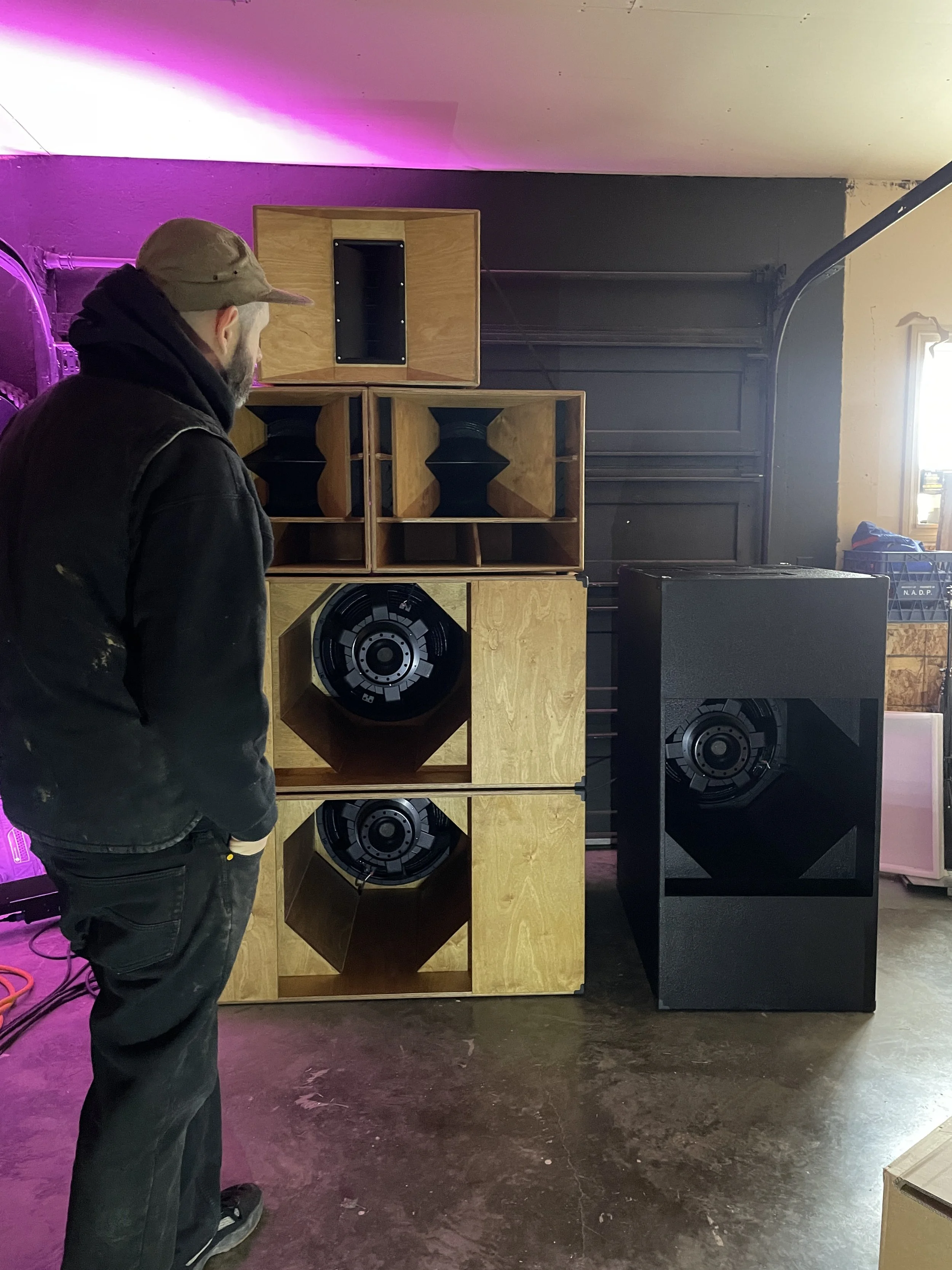 A man in black clothing and a cap stands in front of a stack of wooden speaker cabinets and a large black speaker in a workshop or audio equipment store.