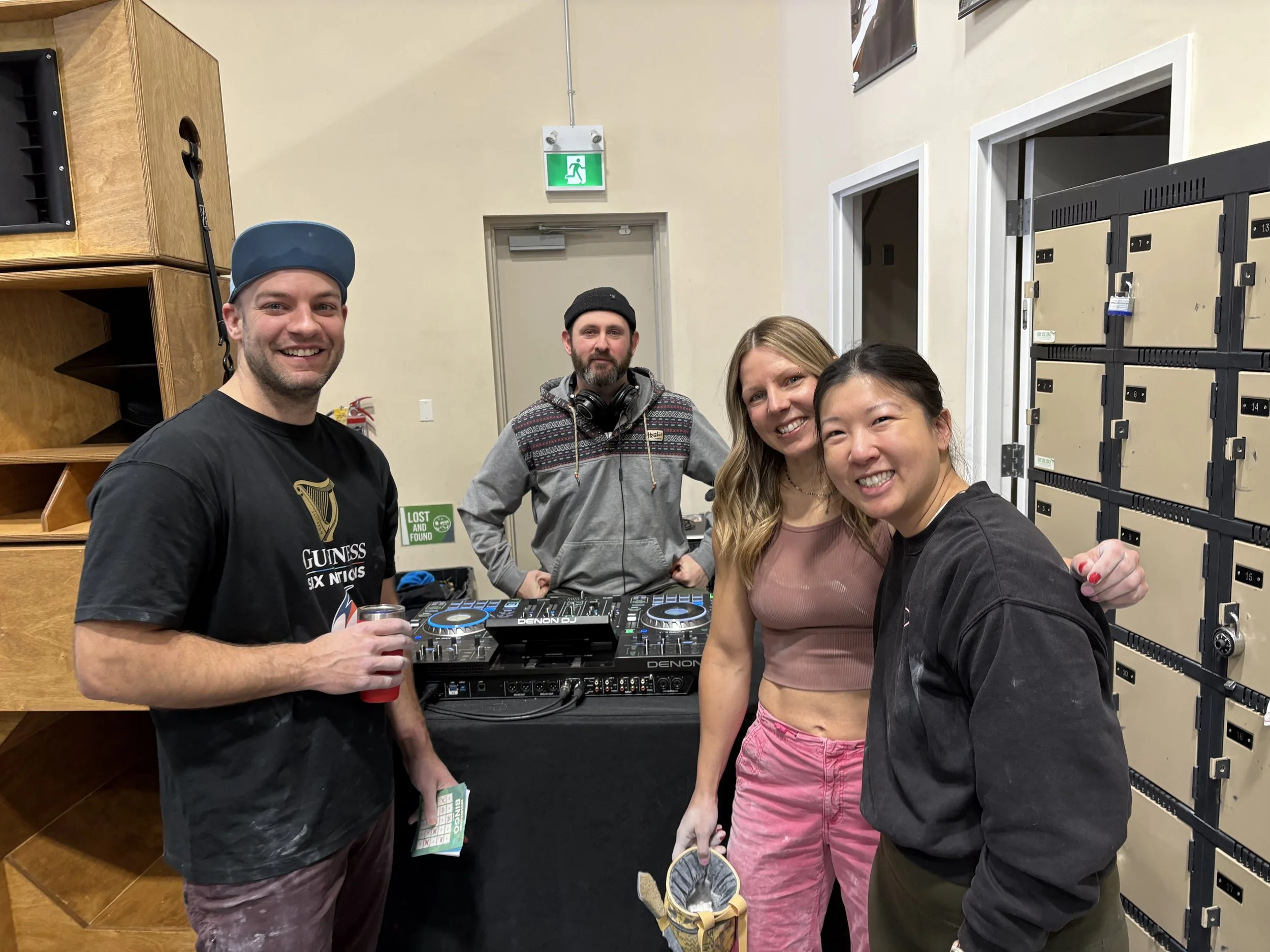 Group of four people at a DJ booth indoors, with two women and two men, smiling at the camera. One man is holding a drink, and the woman in the middle has a basket. The background has lockers and a door with an exit sign.
