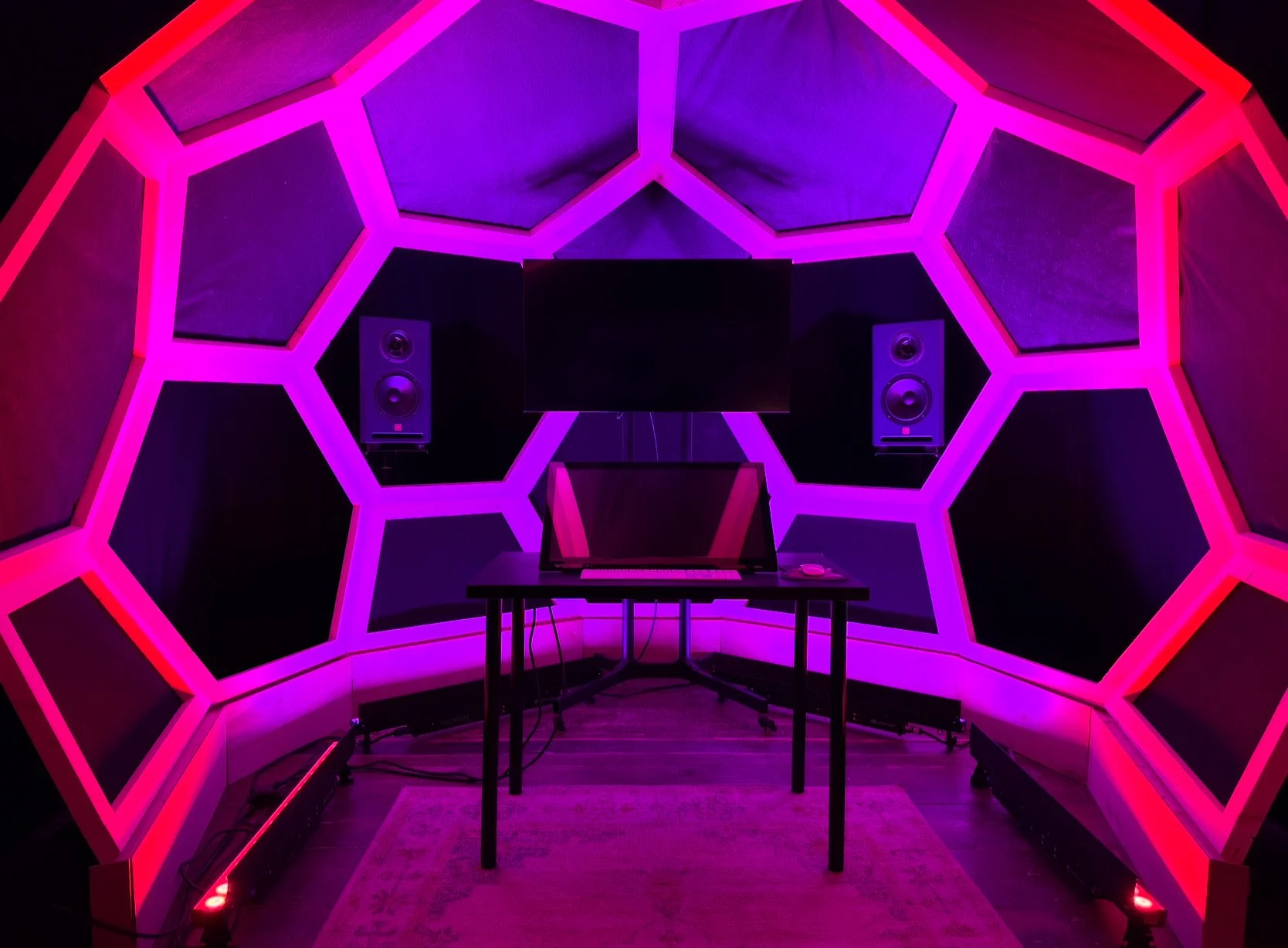 Music studio with a hexagonal lighted wall, computer monitor, studio speakers, and a rug on the floor.