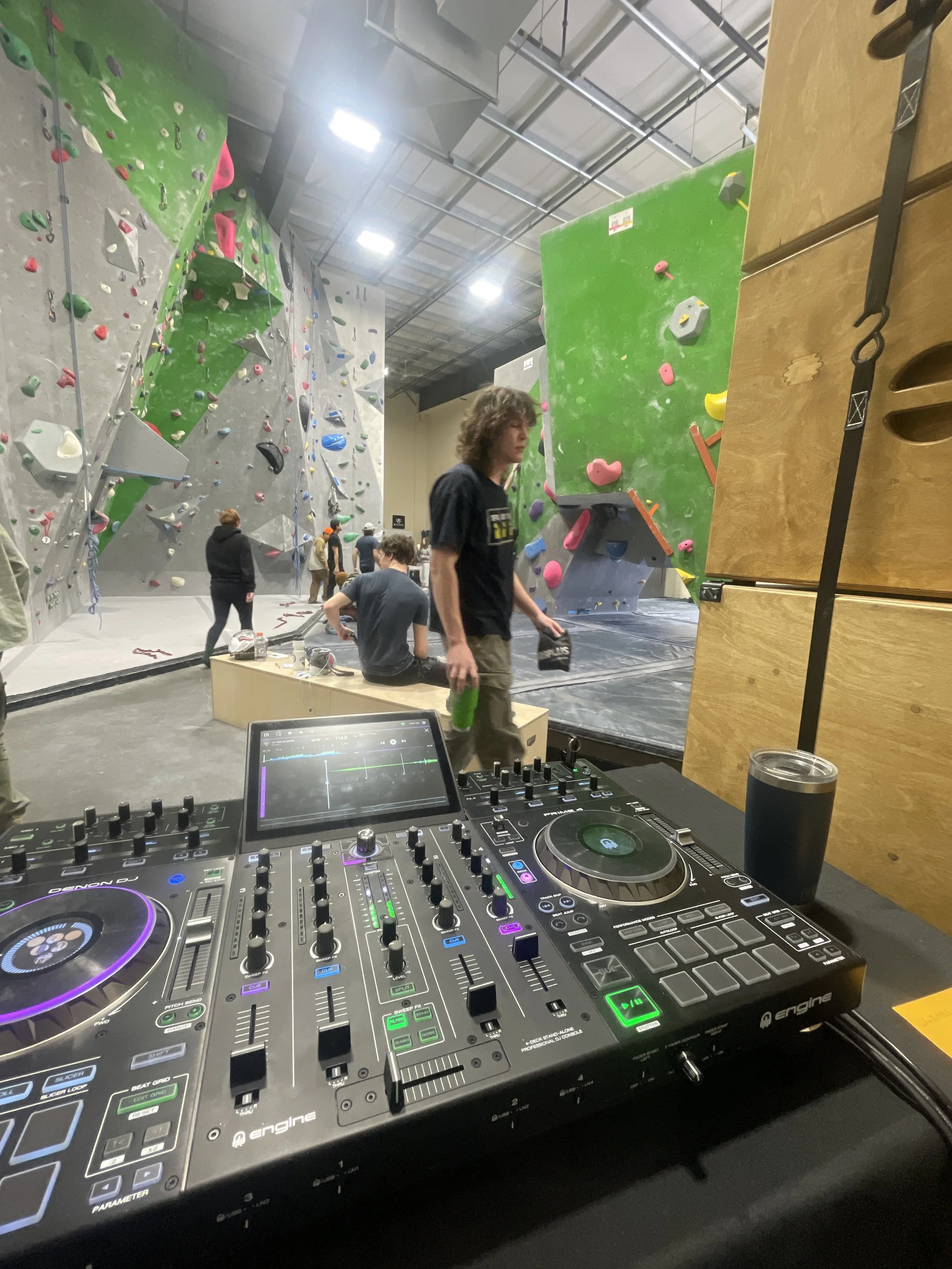 Indoor rock climbing gym with several climbing walls and climbers. A DJ mixer and turntables are visible in the foreground.