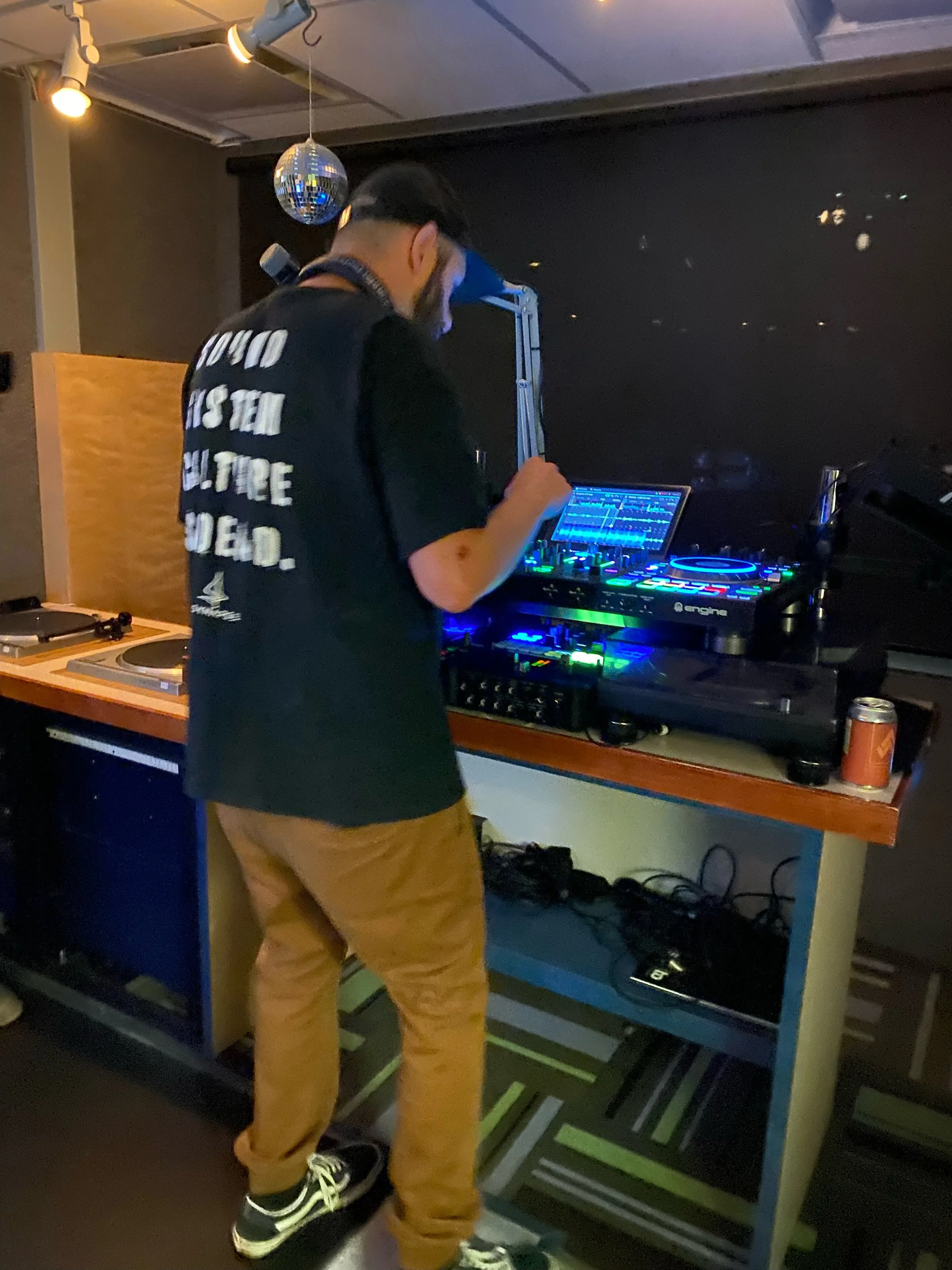 A DJ wearing a black t-shirt with white text, tan pants, and sneakers, working with DJ equipment in a dimly lit room, with a disco ball hanging from the ceiling.