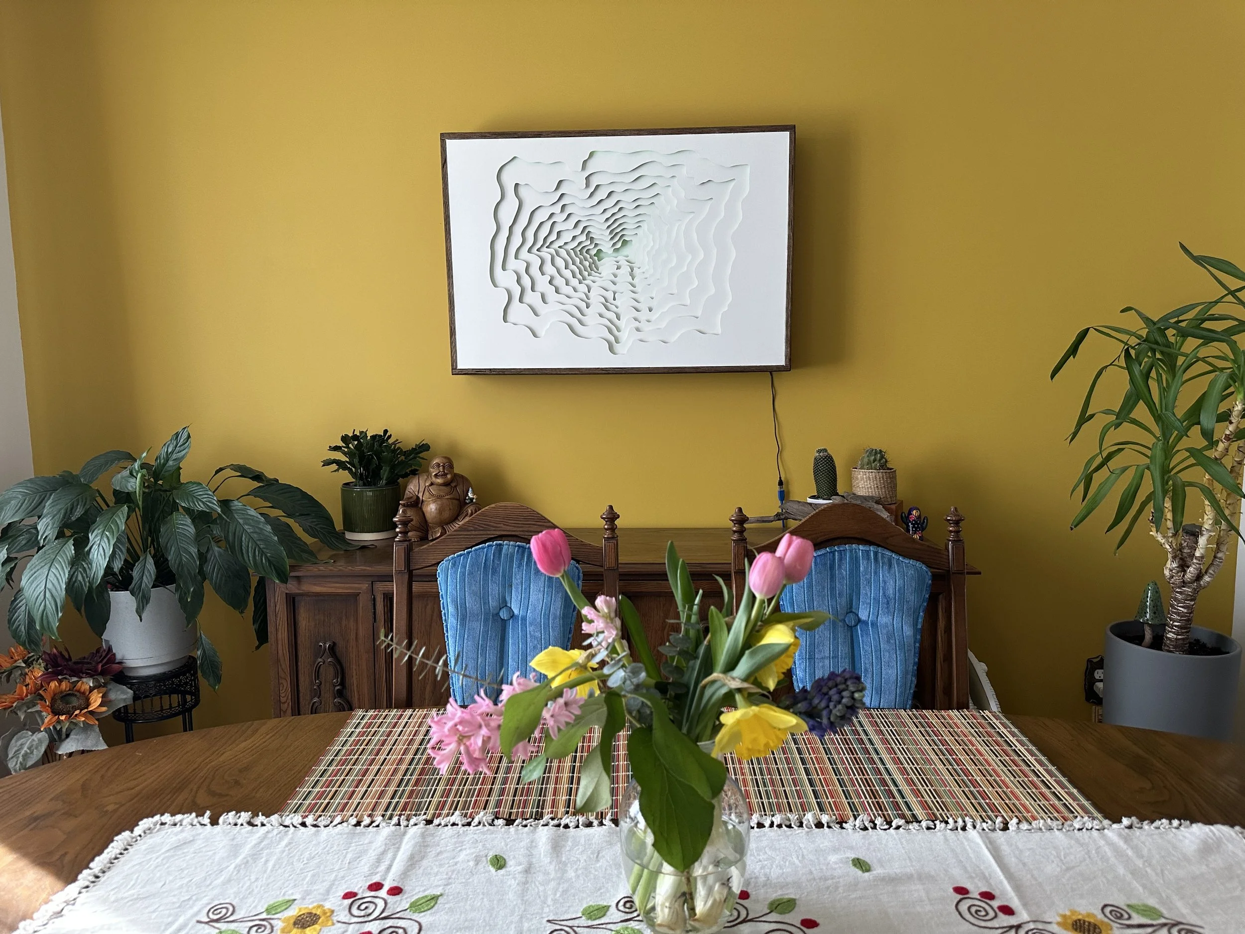 Dining room with a wooden table, a vase with pink and yellow flowers, a yellow wall, a framed white artwork, and plants in the background.