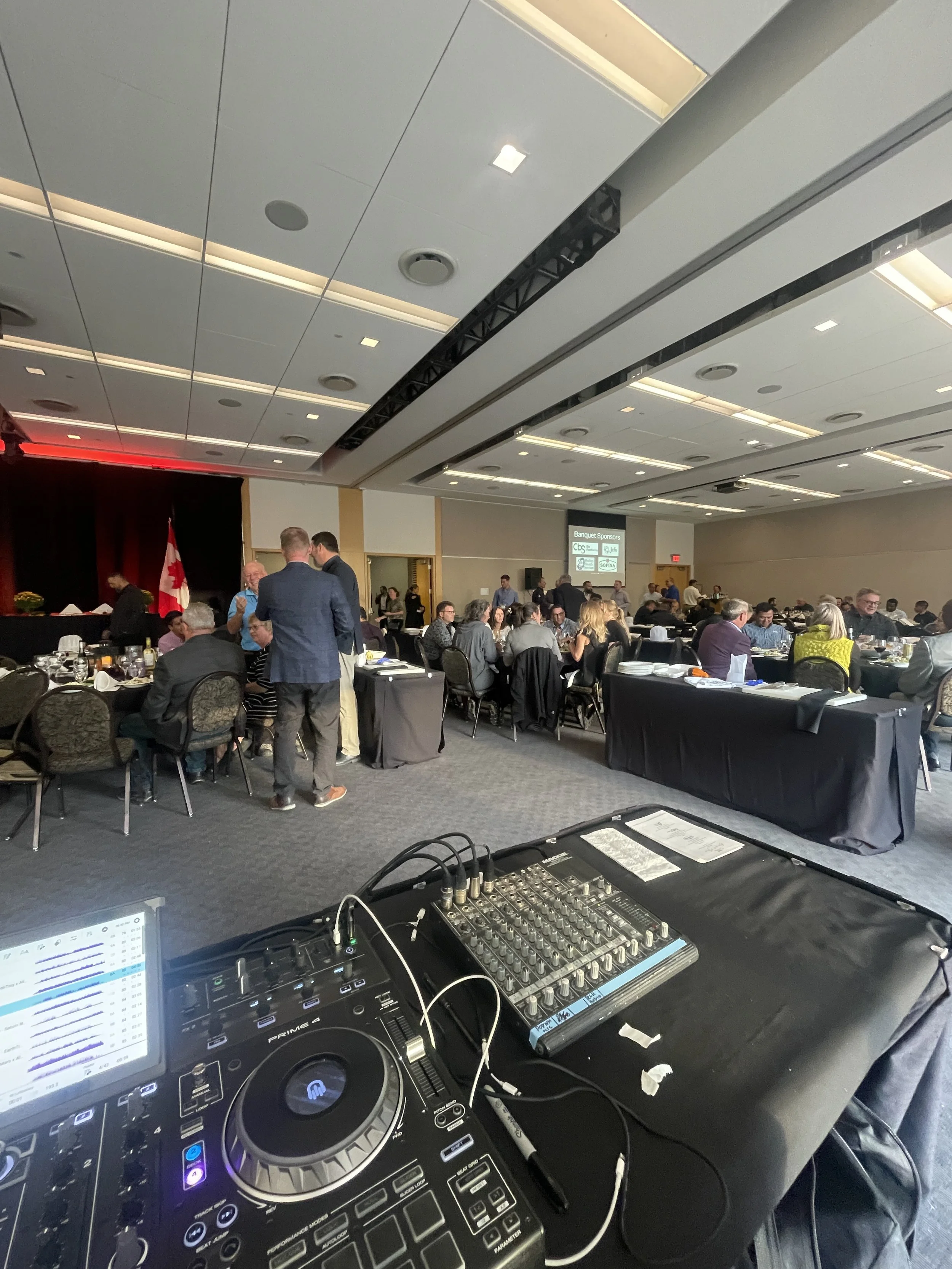 View of a large conference or banquet hall filled with round tables and chairs, many of which are occupied by attendees. In the foreground, there is DJ equipment including a mixer and controller. The room has a high ceiling with recessed lighting, a 