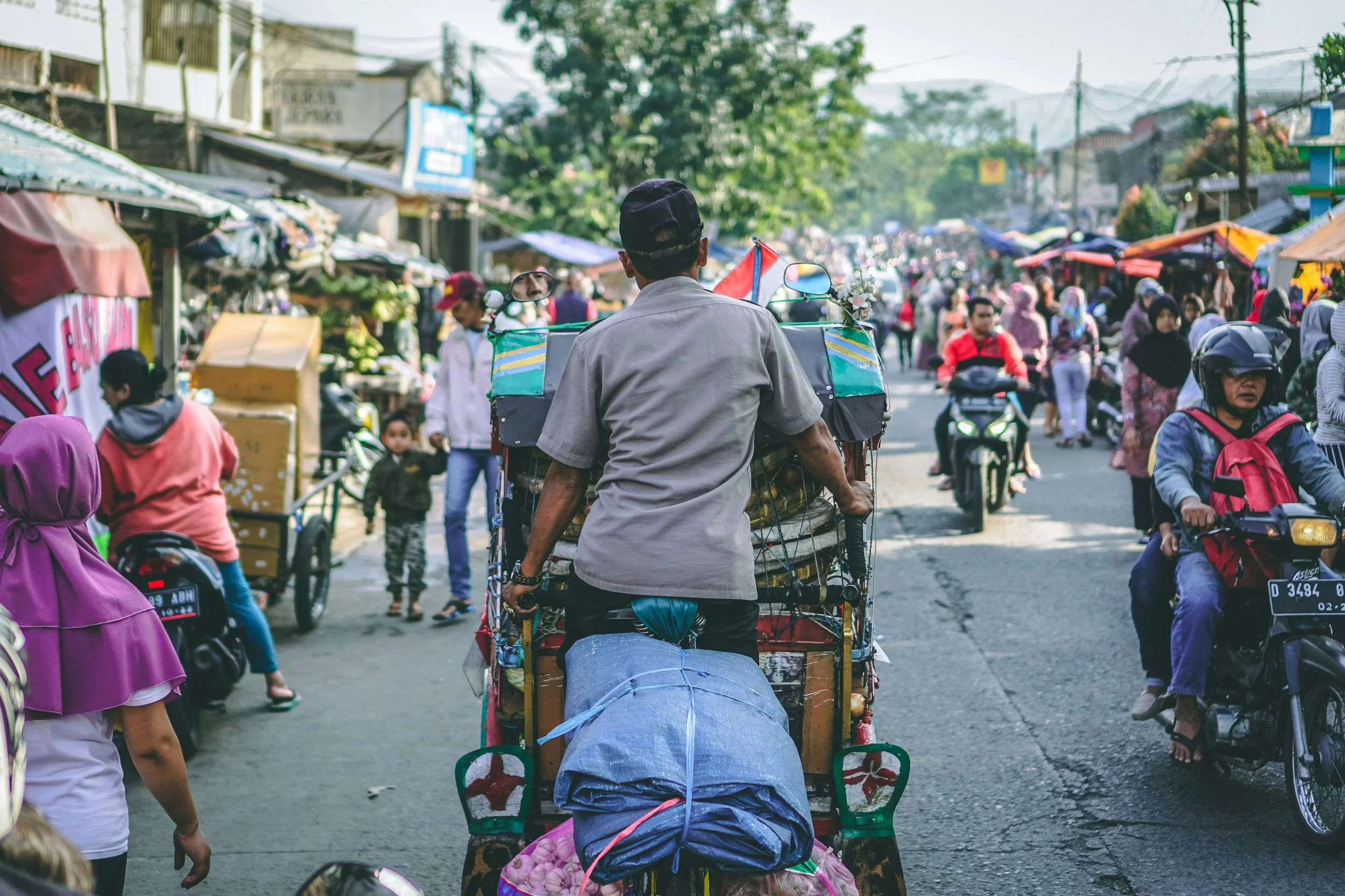 A busy street market scene with people riding motorbikes and carts, some wearing helmets, and a man pushing a rickshaw loaded with goods.