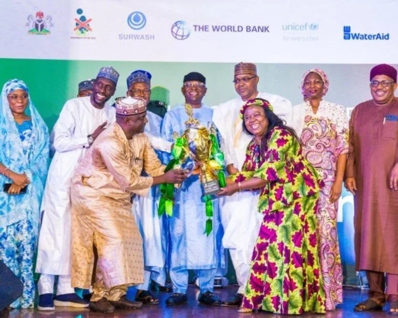 Un groupe d'hommes et de femmes en tenue traditionnelle et en tenue de cérémonie sur scène, recevant un trophée lors d'un événement, avec en arrière-plan des bannières d'organisations telles que la Banque mondiale, l'UNICEF et WaterAid.