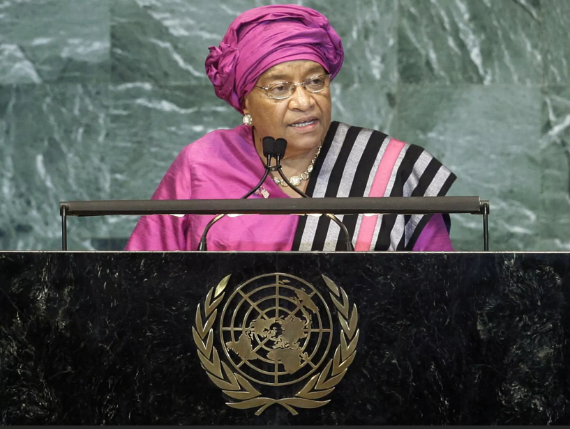 Uma mulher vestida com um traje tradicional cor-de-rosa e um lenço na cabeça, de óculos, a discursar na sede das Nações Unidas, com o emblema da ONU na secretária à sua frente.