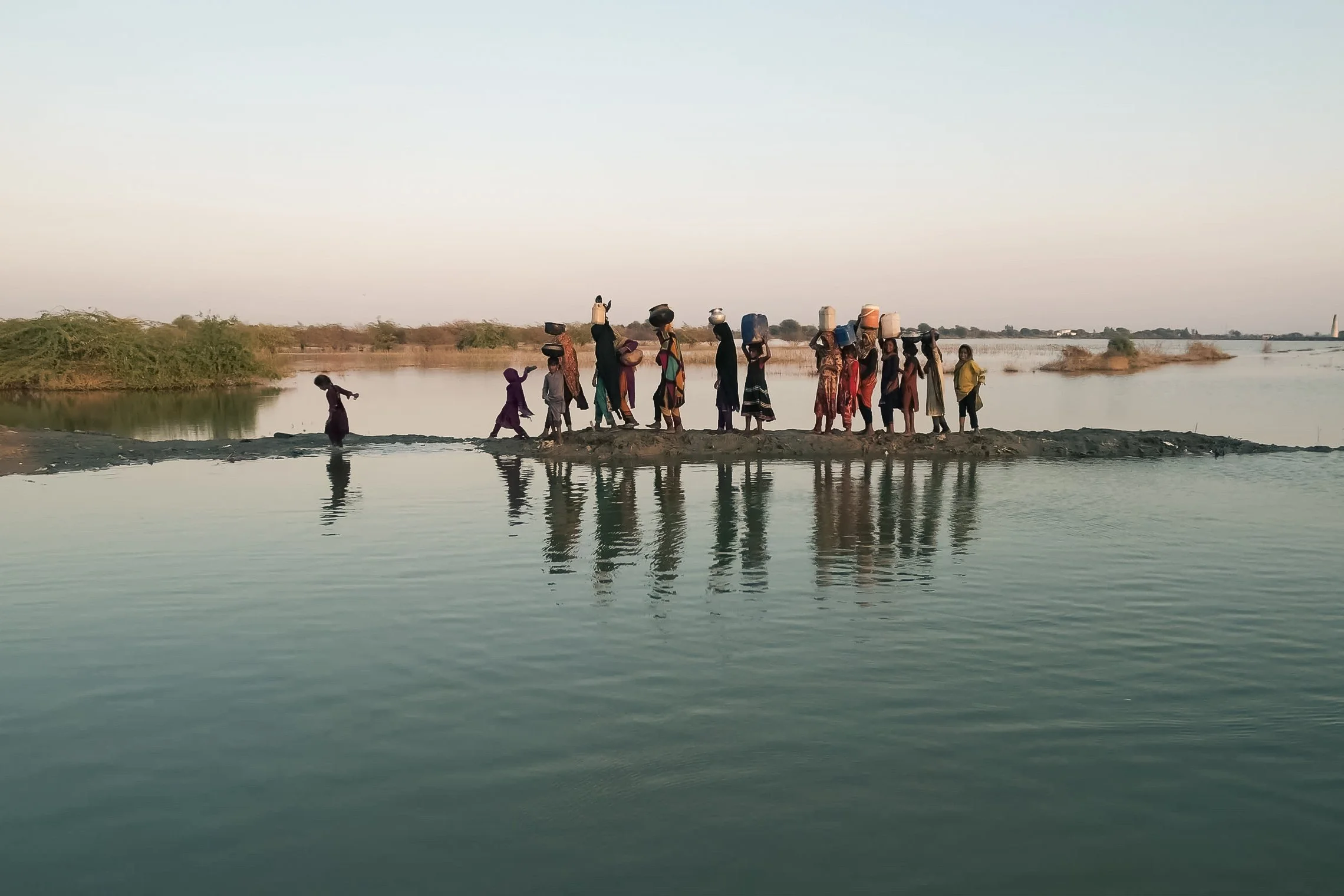 Group of people crossing a river on a small strip of land, with some carrying containers on their heads, at sunset.
