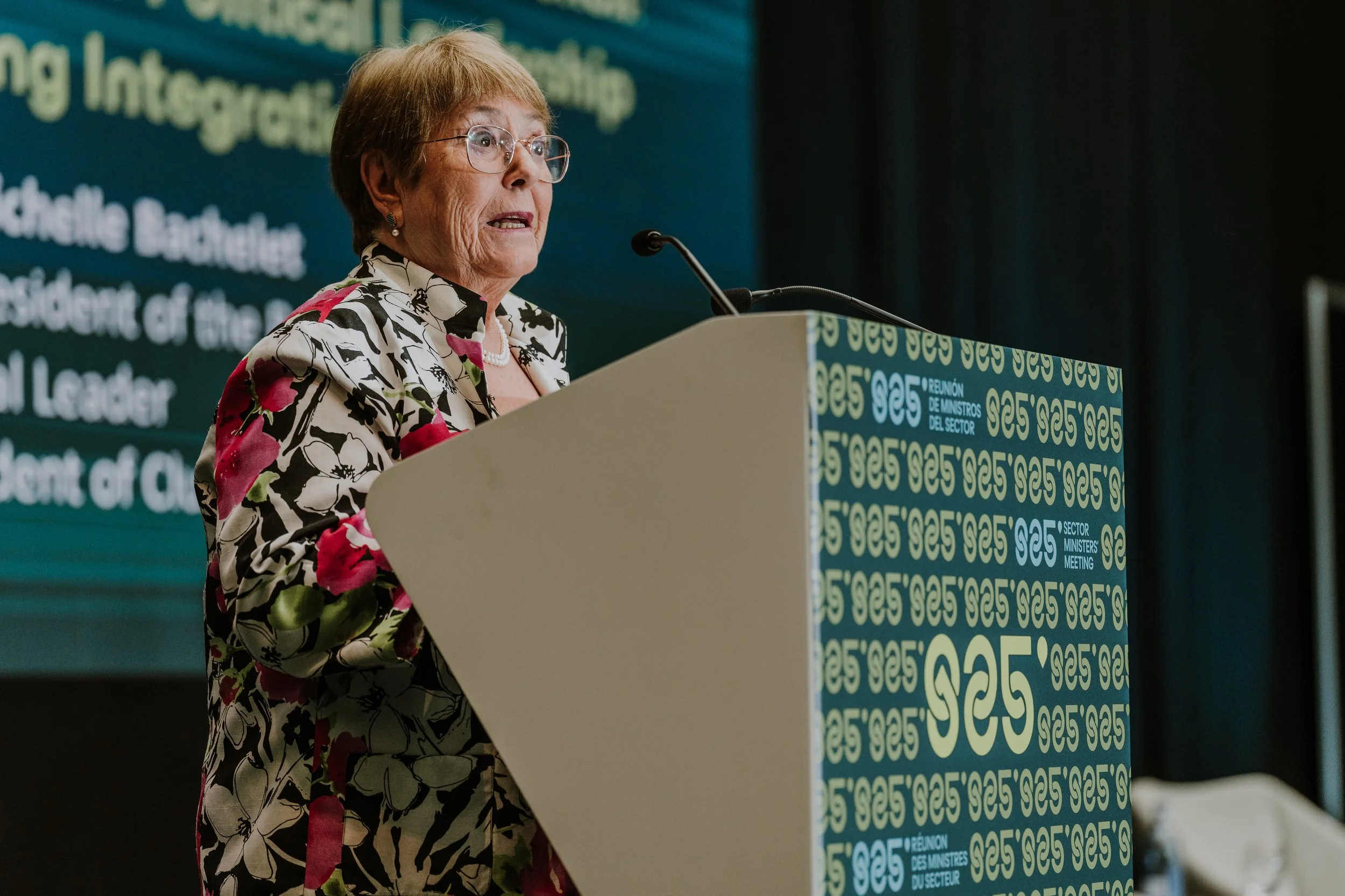 A woman speaking at a podium during a conference, wearing glasses and a floral blazer, with a large screen displaying text behind her.