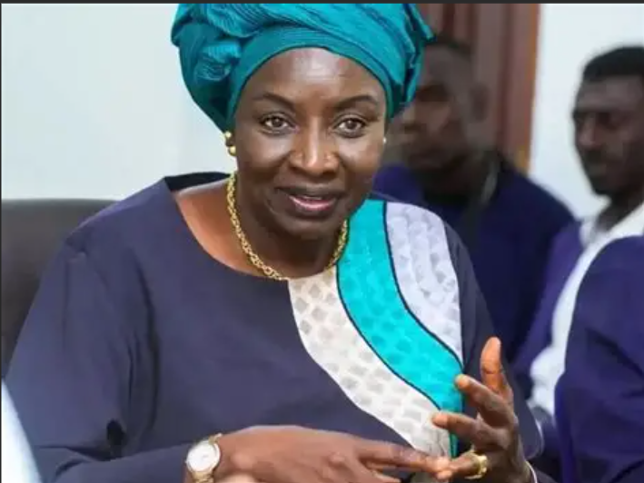 A woman wearing a blue and white traditional outfit with a matching headwrap, gold jewelry, and a wristwatch, gesturing with her hands and speaking, with other people in the background.