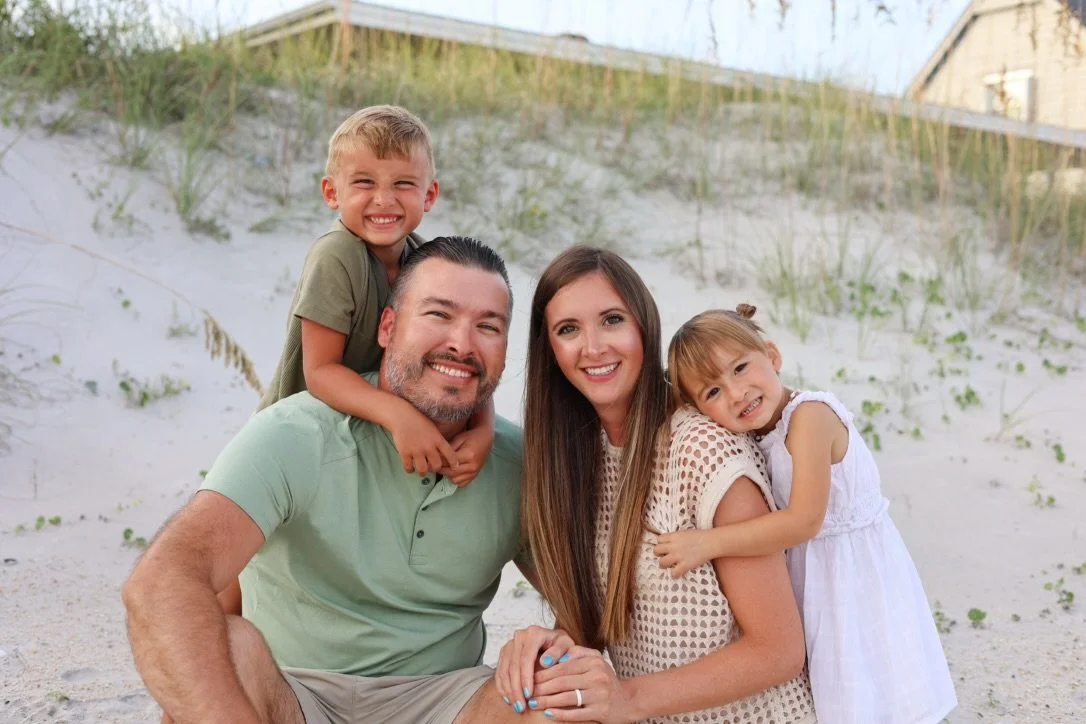 Family photo of a man, woman, a young boy, and a young girl smiling and posing on a sandy beach with grassy dunes in the background