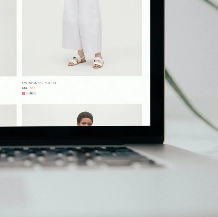 Laptop screen displaying a shopping website with a white dress, white sandals, and text about a round-neck T-shirt.