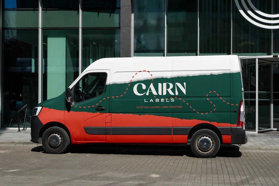 A delivery van parked in front of a modern building with glass windows, featuring a colorful wrap design with green, orange, and white colors, and the words "Cairn Labels" and "Scottish Luxury Label Printers" on the side.