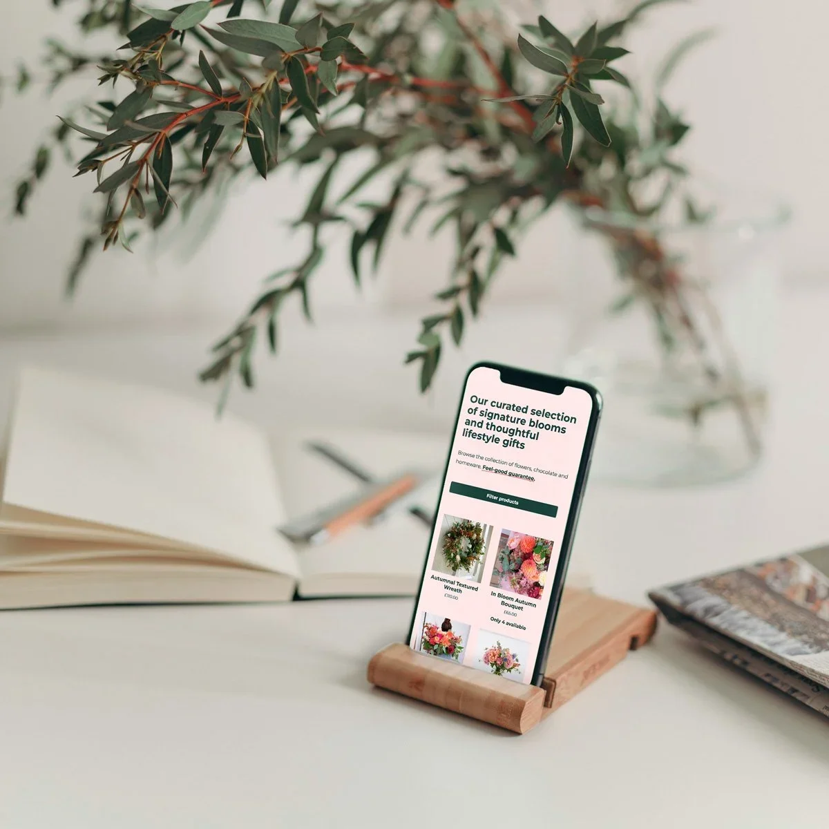 Smartphone on a wooden stand displaying an online flower shop website. In the background, there is a leafy plant in a glass vase, an open book, and a pen on a white surface.