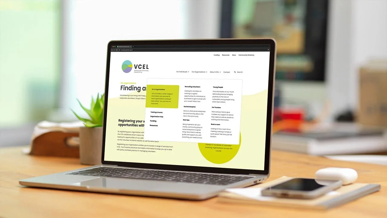Open laptop on a wooden desk displaying a website for VCEL with content about volunteering and organization opportunities. Nearby are a plant in a gray pot, a white wireless mouse, a smartphone, and a notepad with a pen.