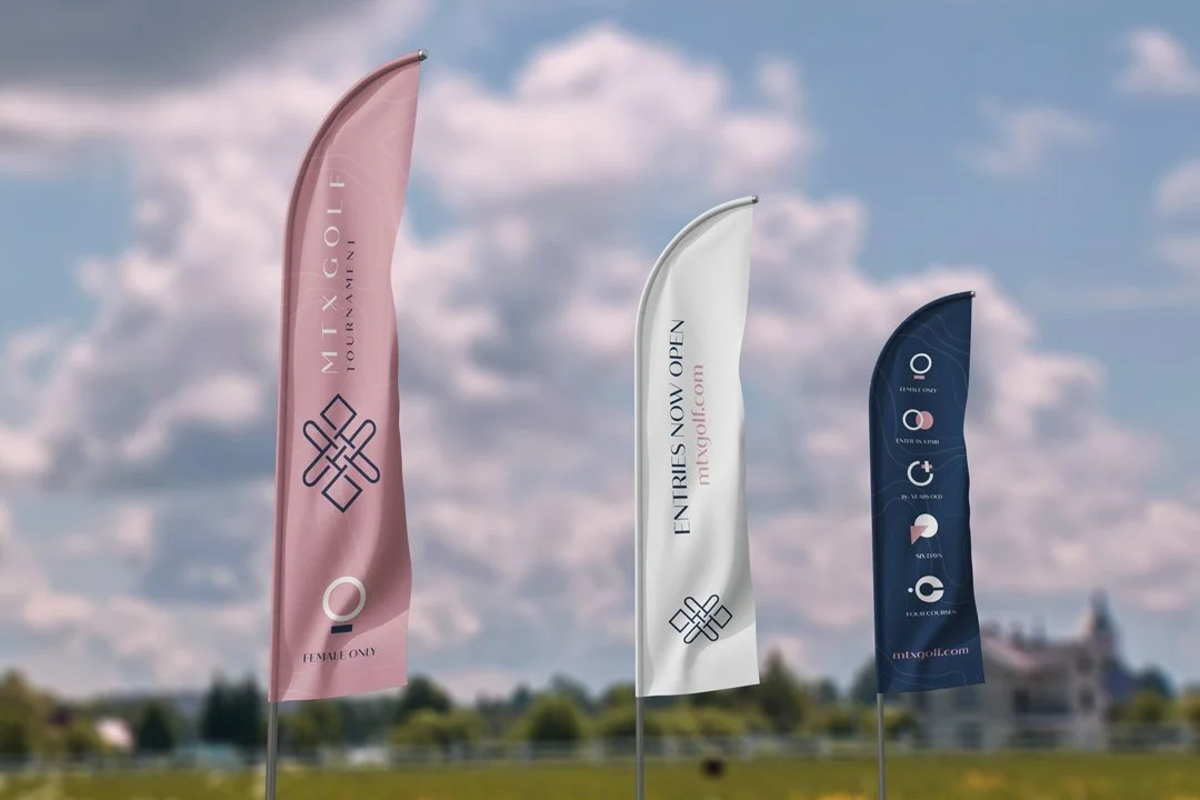 Three vertical banners on poles outdoors, with a grassy background and cloudy sky. The banners are pink, white, and navy blue, displaying logos and text for a golf female-only event.