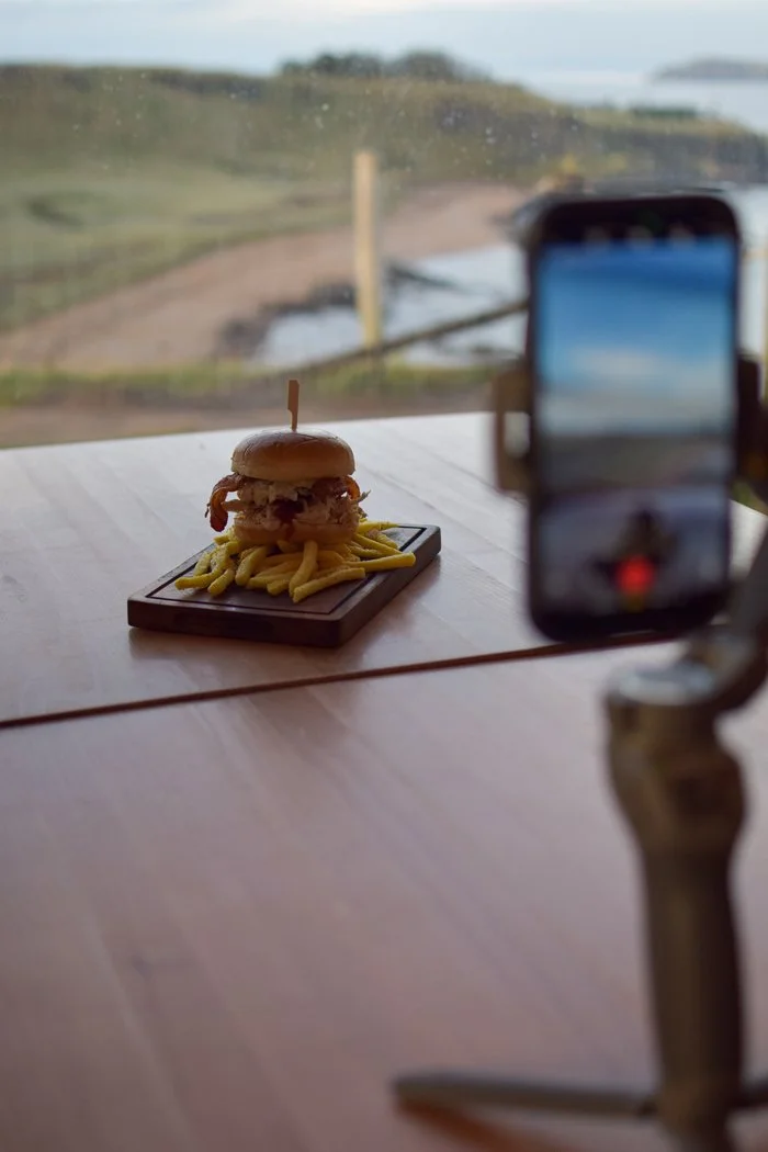 A burger with fries on a wooden tray, placed on a table near a window with a countryside view, being prepared for photo shoot using a smartphone on a tripod.
