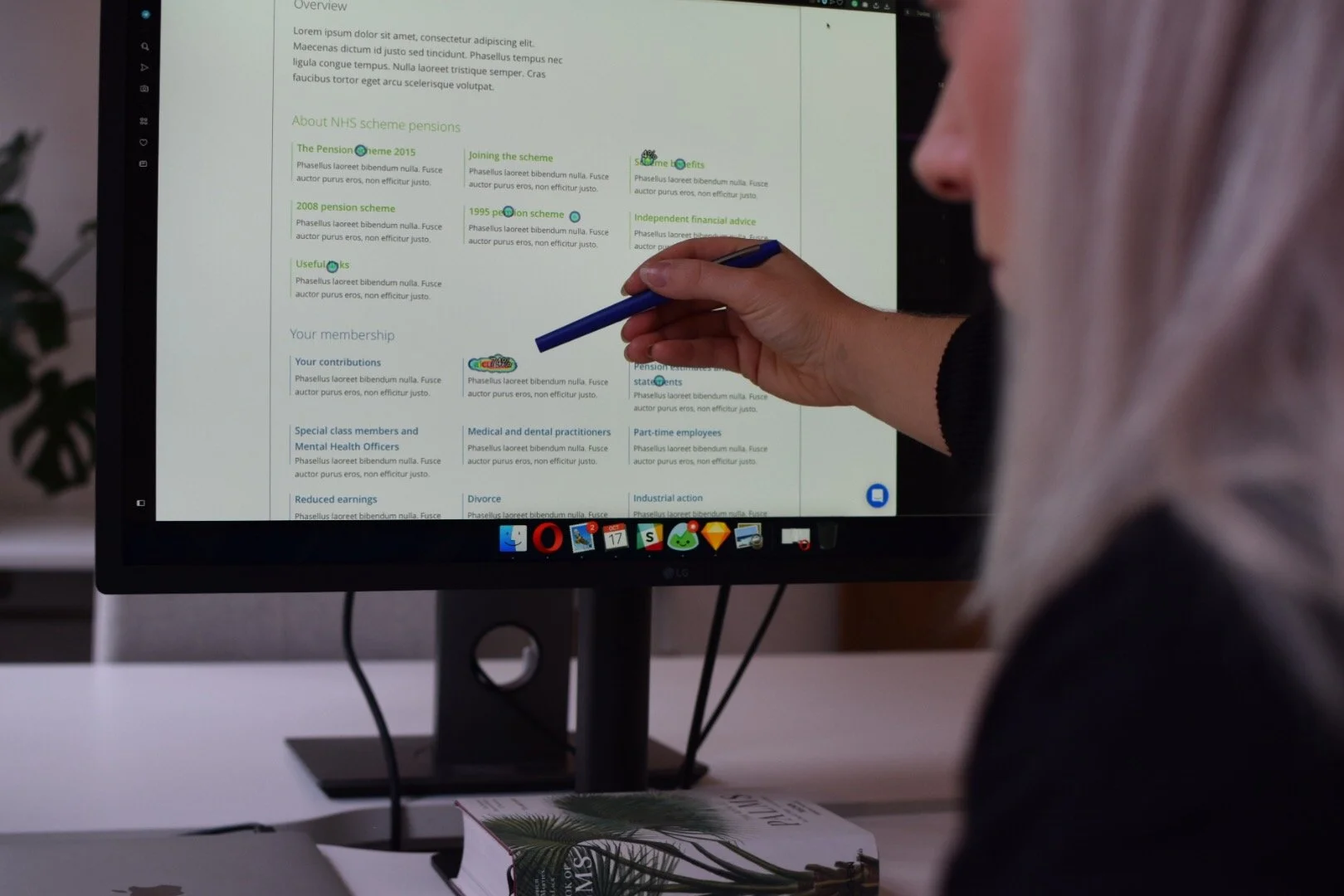 A person pointing at a computer monitor displaying a document with text and highlighted sections related to NHS scheme pensions. The monitor is on a desk with a closed book and a laptop in front, and the person's face is partially visible on the right side of the image.