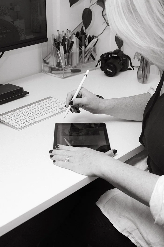 Woman using a stylus on a tablet at a desk with a keyboard, camera, and office supplies, in black and white.