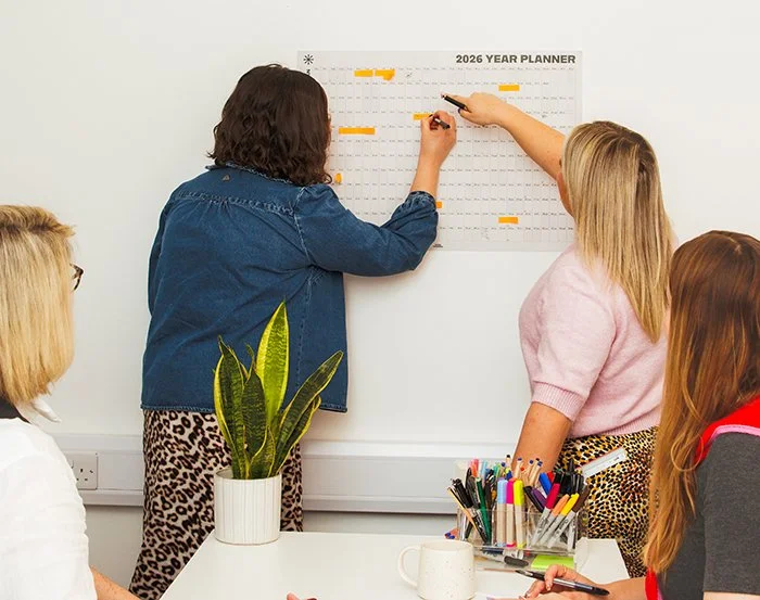 Four women sitting around a table with office supplies, one woman pointing at a 2026 year planner on the wall, and two women standing and discussing it.