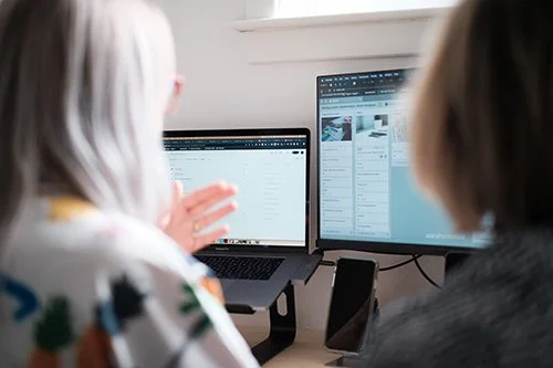 Two people working at a desk with one laptop and one monitor, displaying spreadsheets or data sheets.