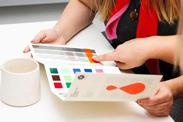 Person holding a color swatch and a brochure with a red fish logo, sitting at a white table next to a white mug.