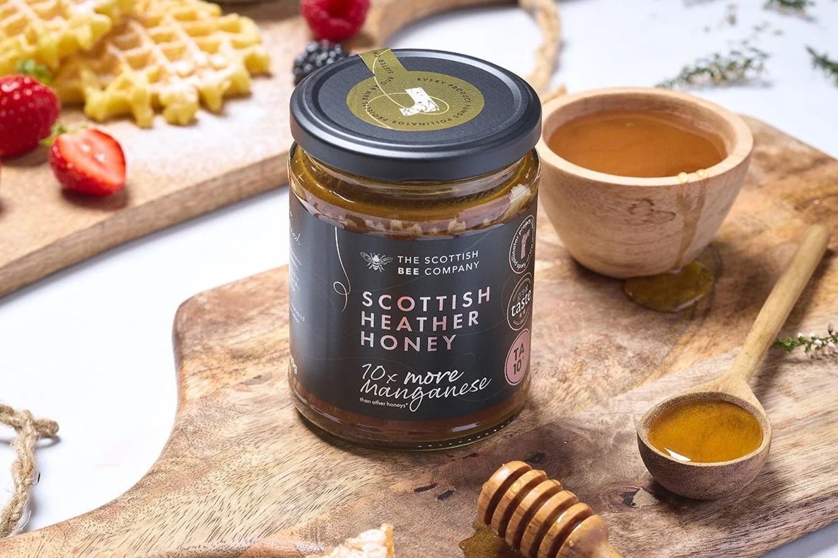 A jar of Scottish Heather Honey on a wooden board with a honey dipper and a small bowl of honey nearby, strawberries and a waffle cake in the background.