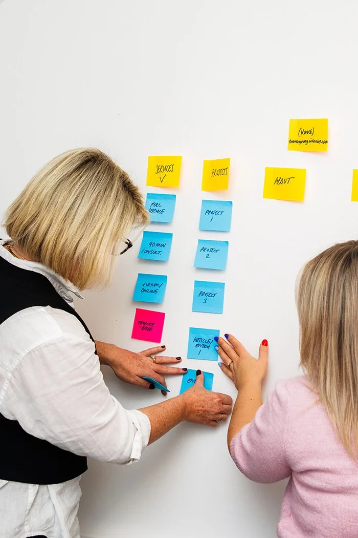 Two women working on a project plan with colorful sticky notes on a whiteboard.