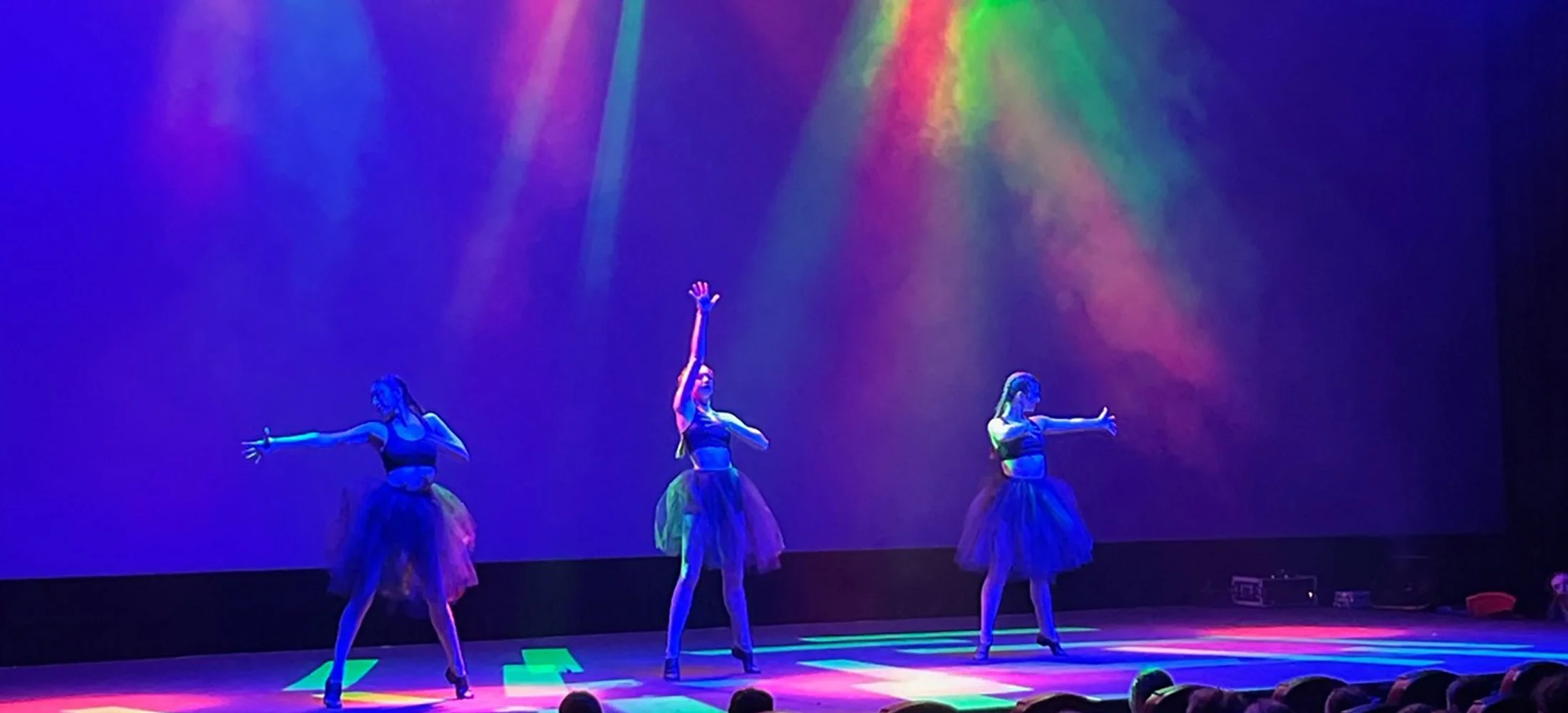 Three female dancers performing on stage at the Double E VT under colorful lights, wearing black tops and tutus.