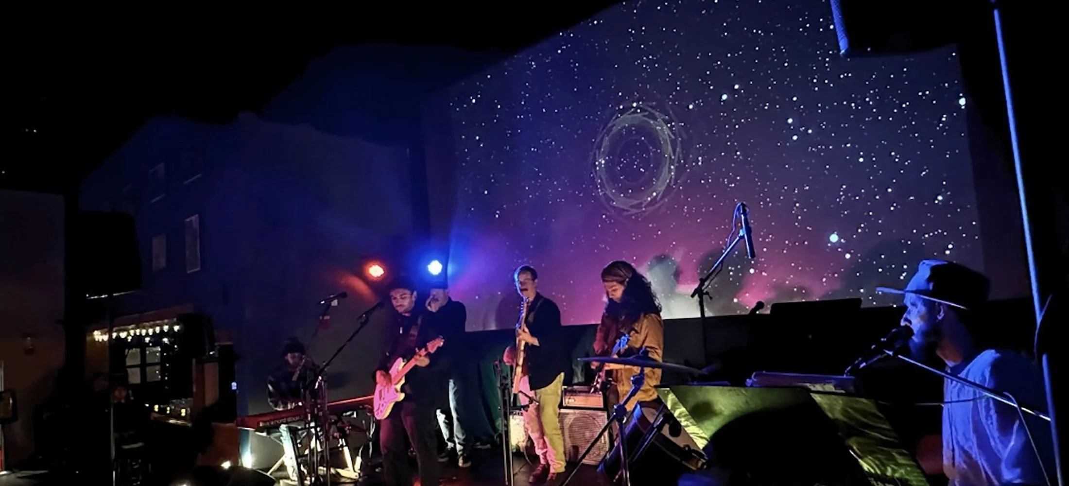 Live band performing on stage at the Double E VT with cosmic starfield projection in the background, featuring multiple musicians playing instruments.