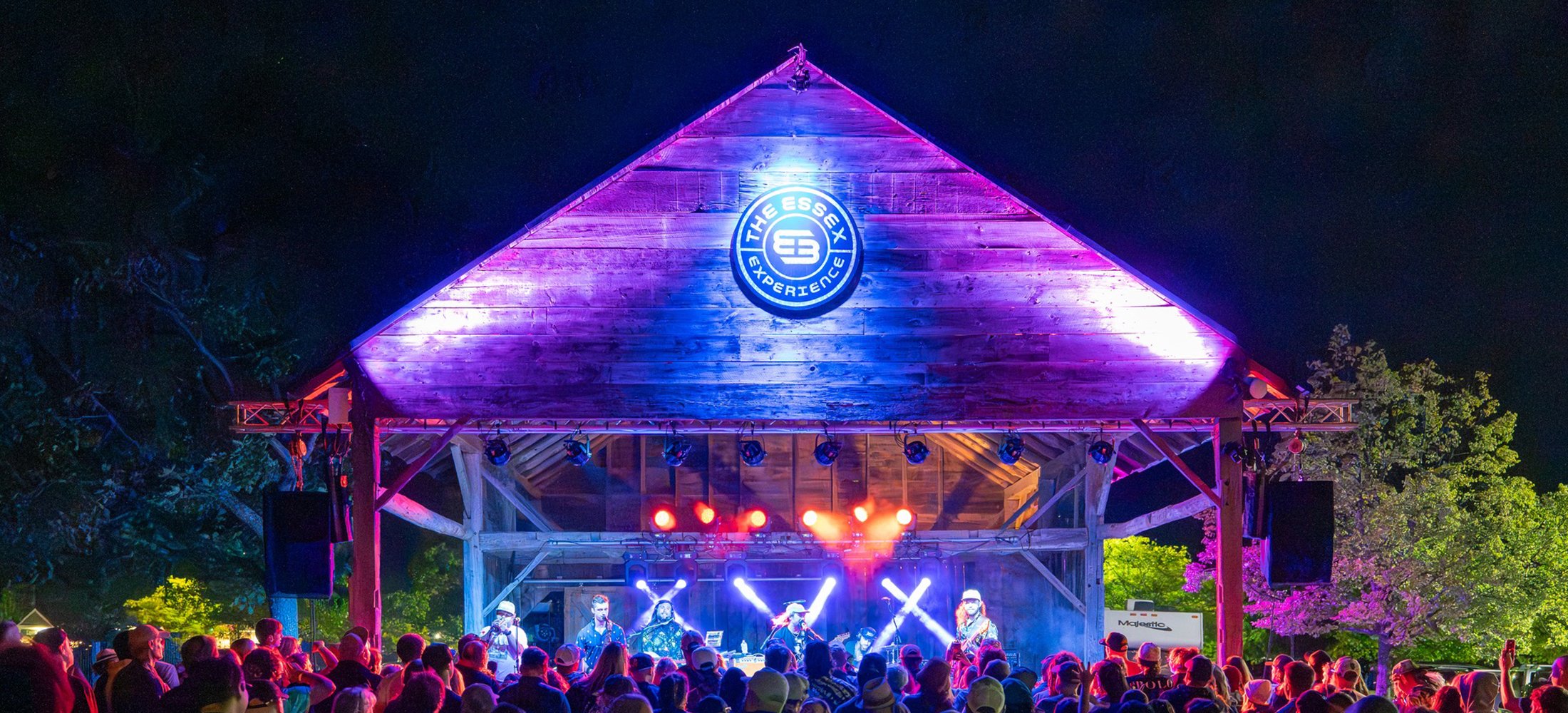 Outdoor music concert at night at the Double E VT with a wooden stage illuminated by colorful lights, surrounded by an audience and trees with pink blossoms.