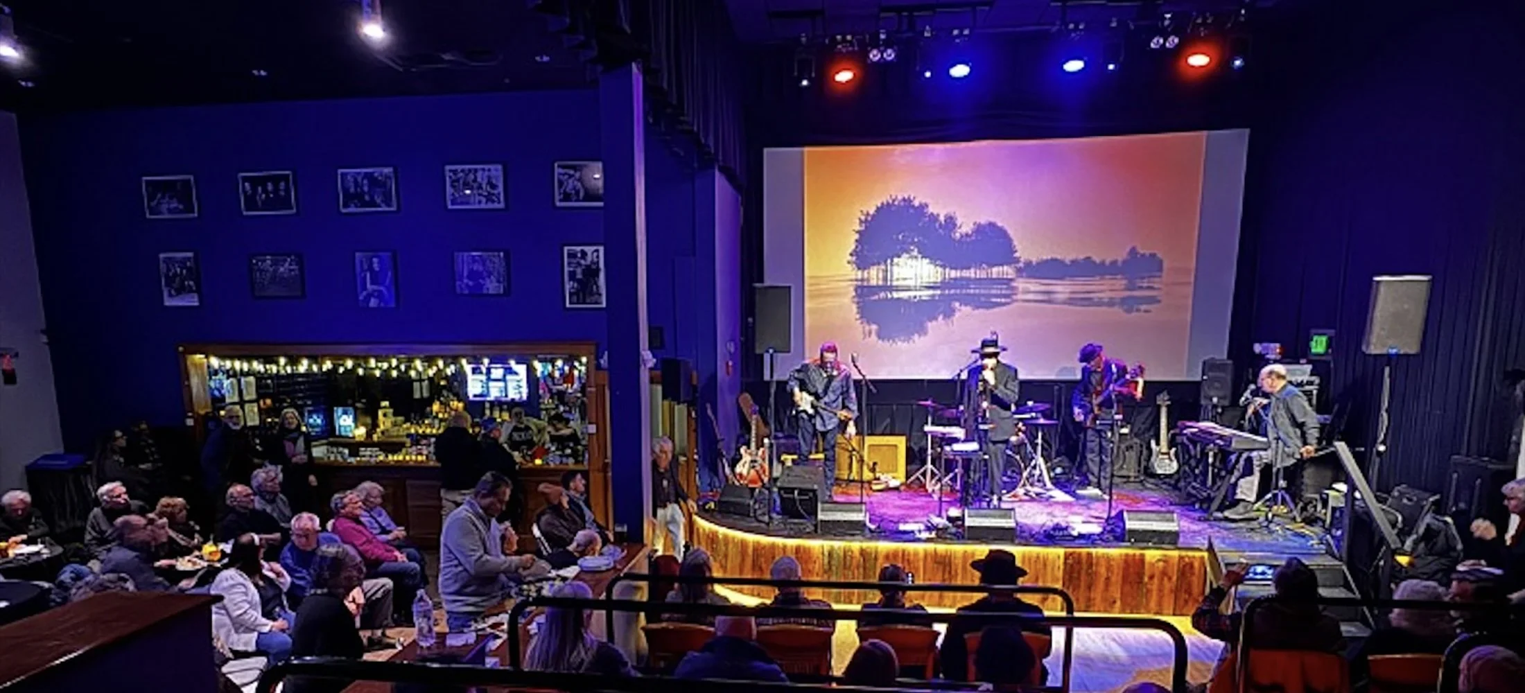 A live music performance on a stage at the Double E VT with a band playing instruments including guitars and drums, a large screen showing a sunset scene with trees, and an audience seated at tables and in chairs in a dark indoor venue.
