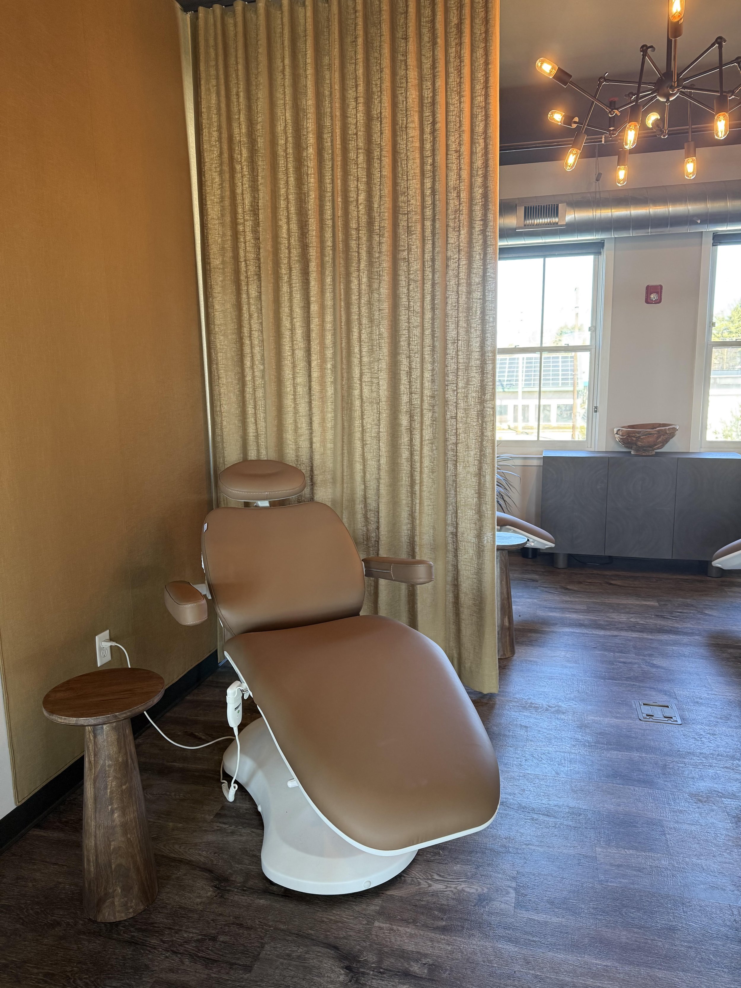 A medical examination chair next to a small circular wooden side table in a modern room with wood floors, gold curtains, and large windows, decorated with a black metal chandelier.