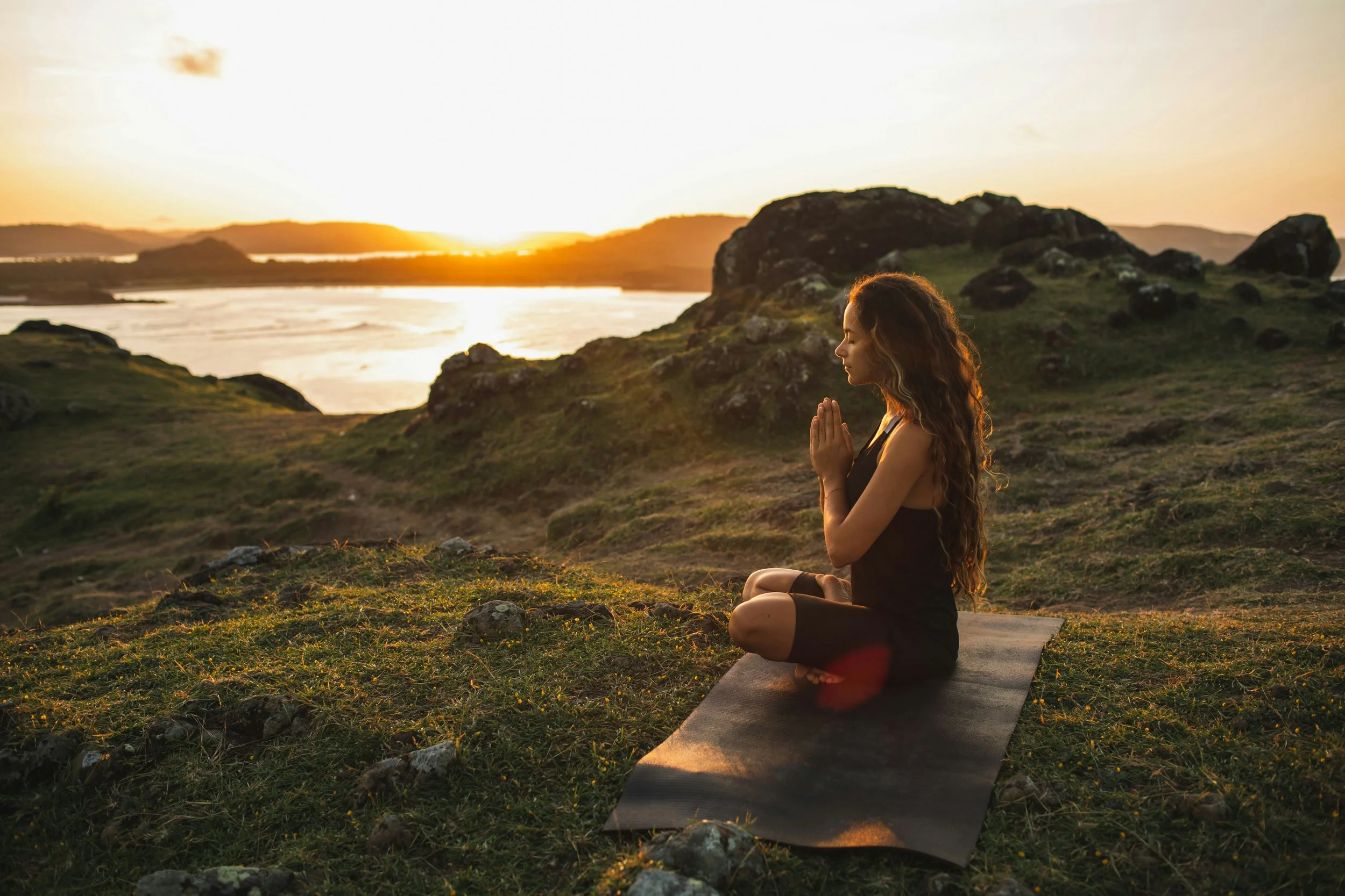 A woman practicing yoga outdoors on a mat near a body of water during sunset, sitting cross-legged with hands in prayer position.