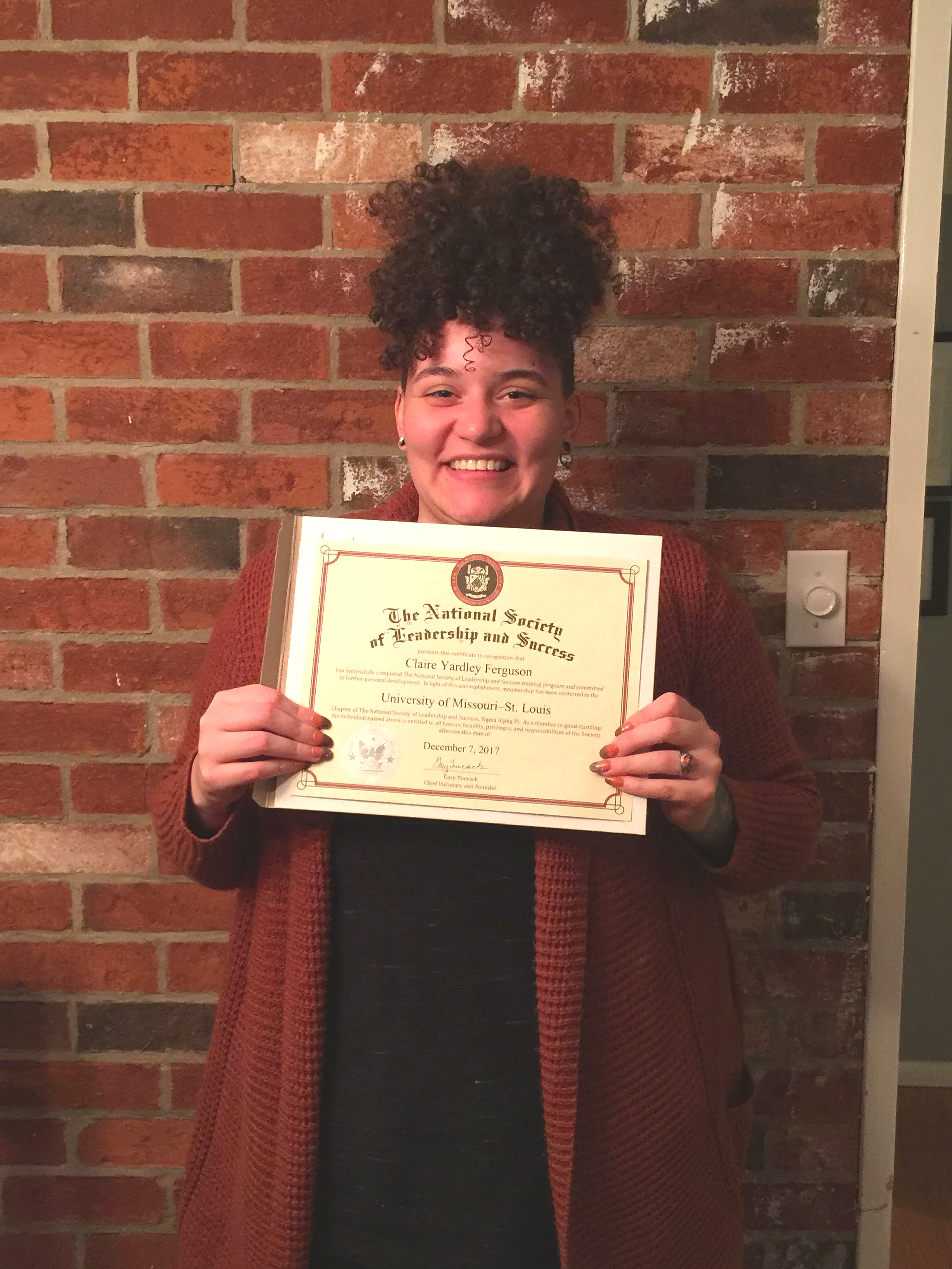 Claire Ferguson smiling and holding a certificate in front of a brick wall