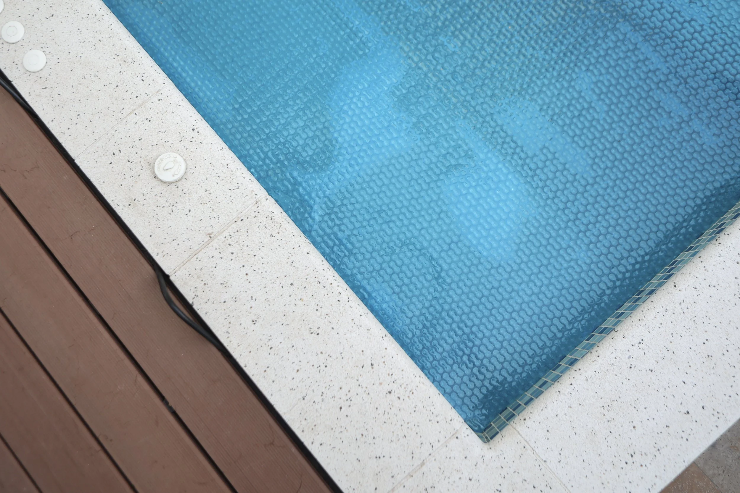 Close-up of a corner of a swimming pool with blue water, surrounding white textured tiles, and a wooden deck.